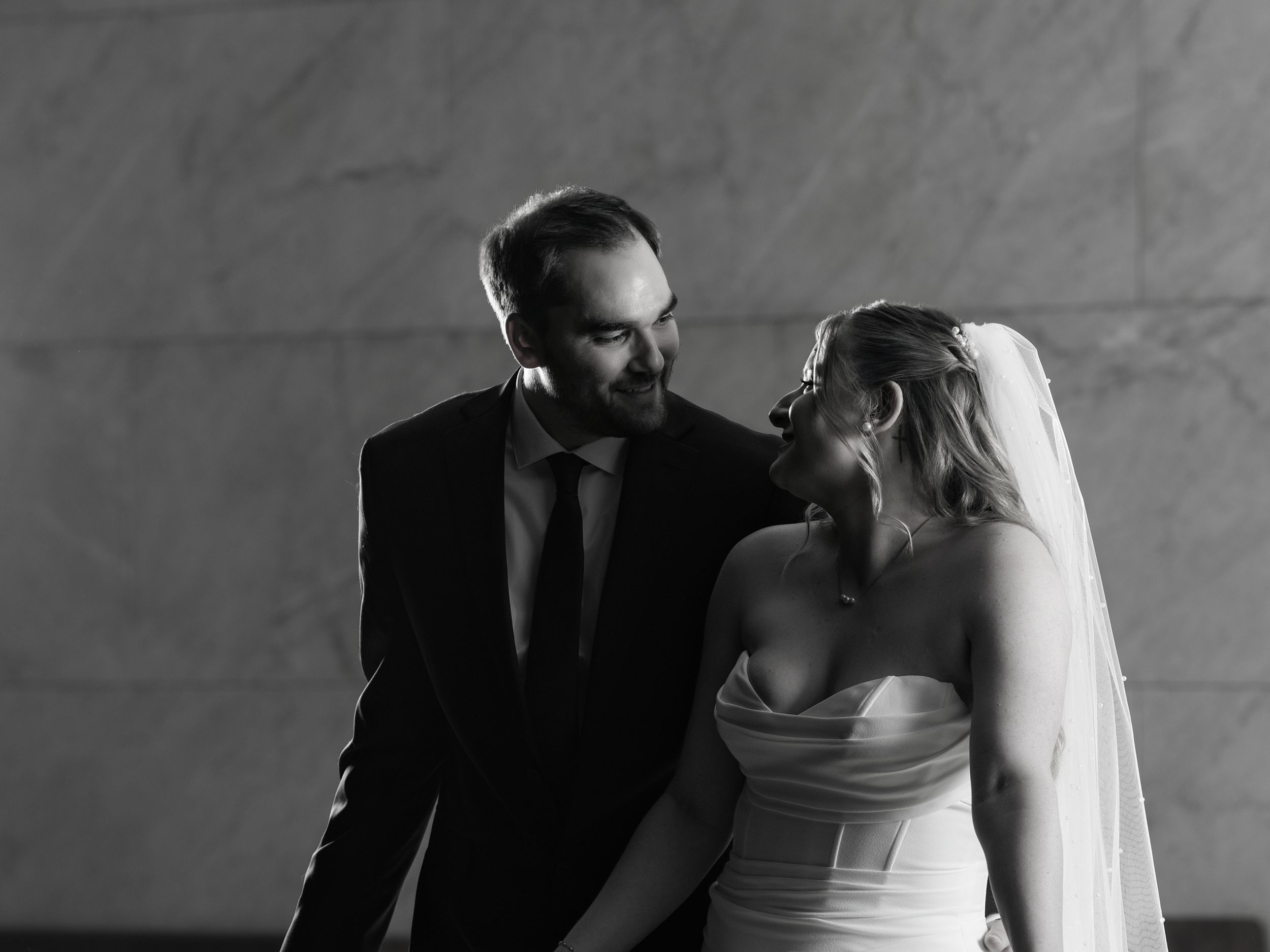 A bride and groom laugh together against a marble wall in black and white