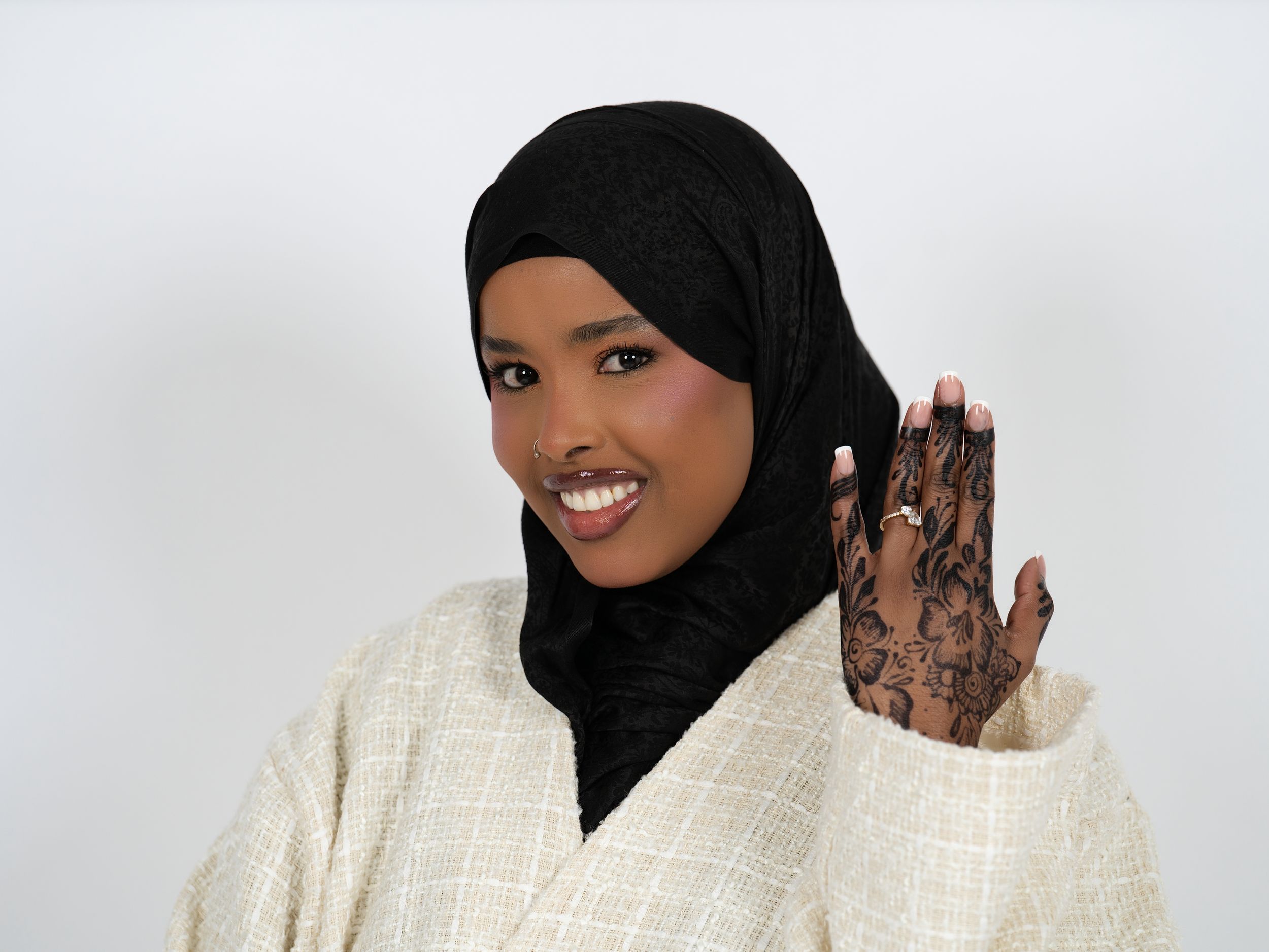A smiling woman in a black headscarf raising a henna-patterned hand against a light studio backdrop