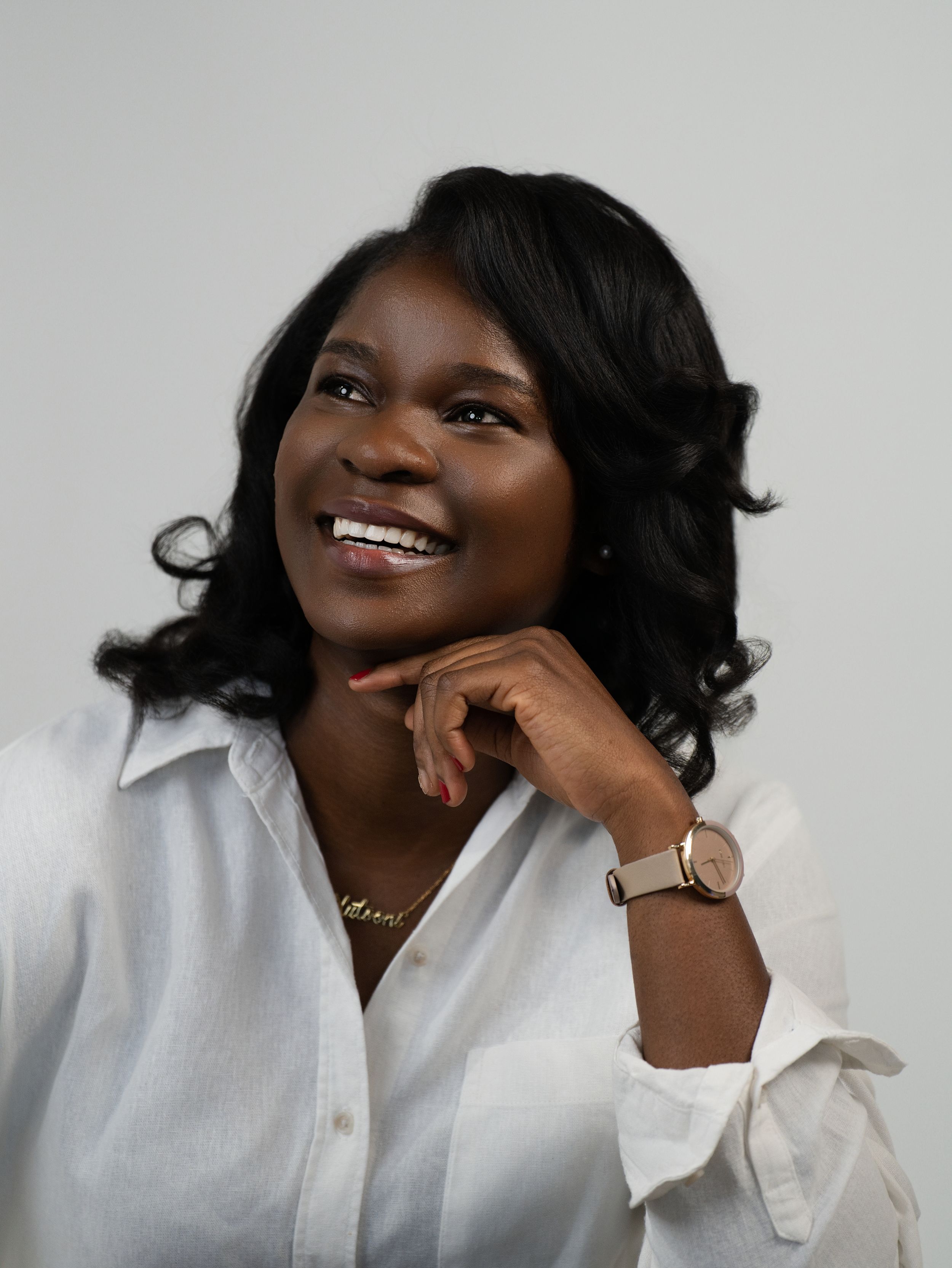 A woman in a white blouse smiles with her hand beneath her chin in a studio