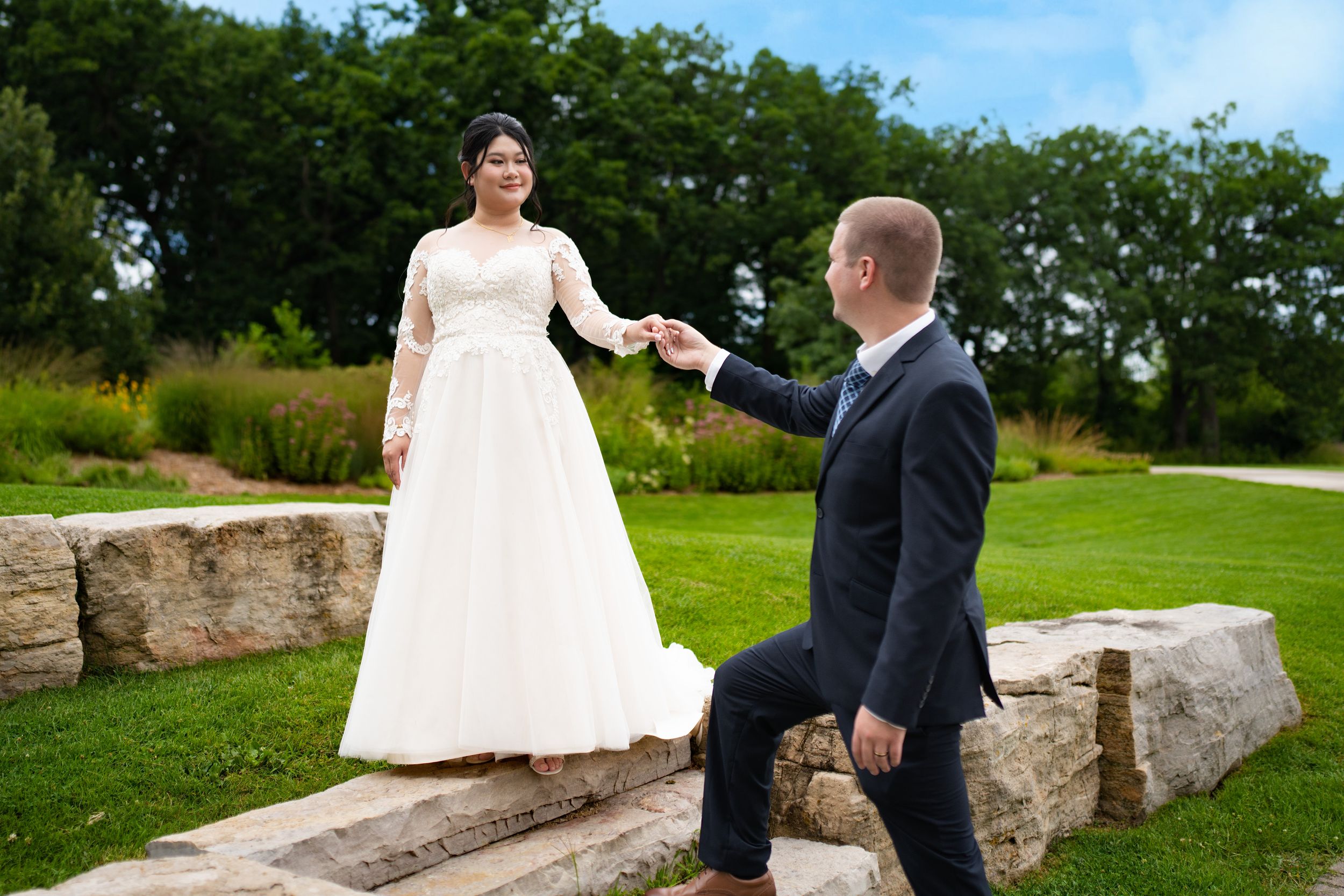 A groom holds the bride's hand as she steps along a stone wall in a garden