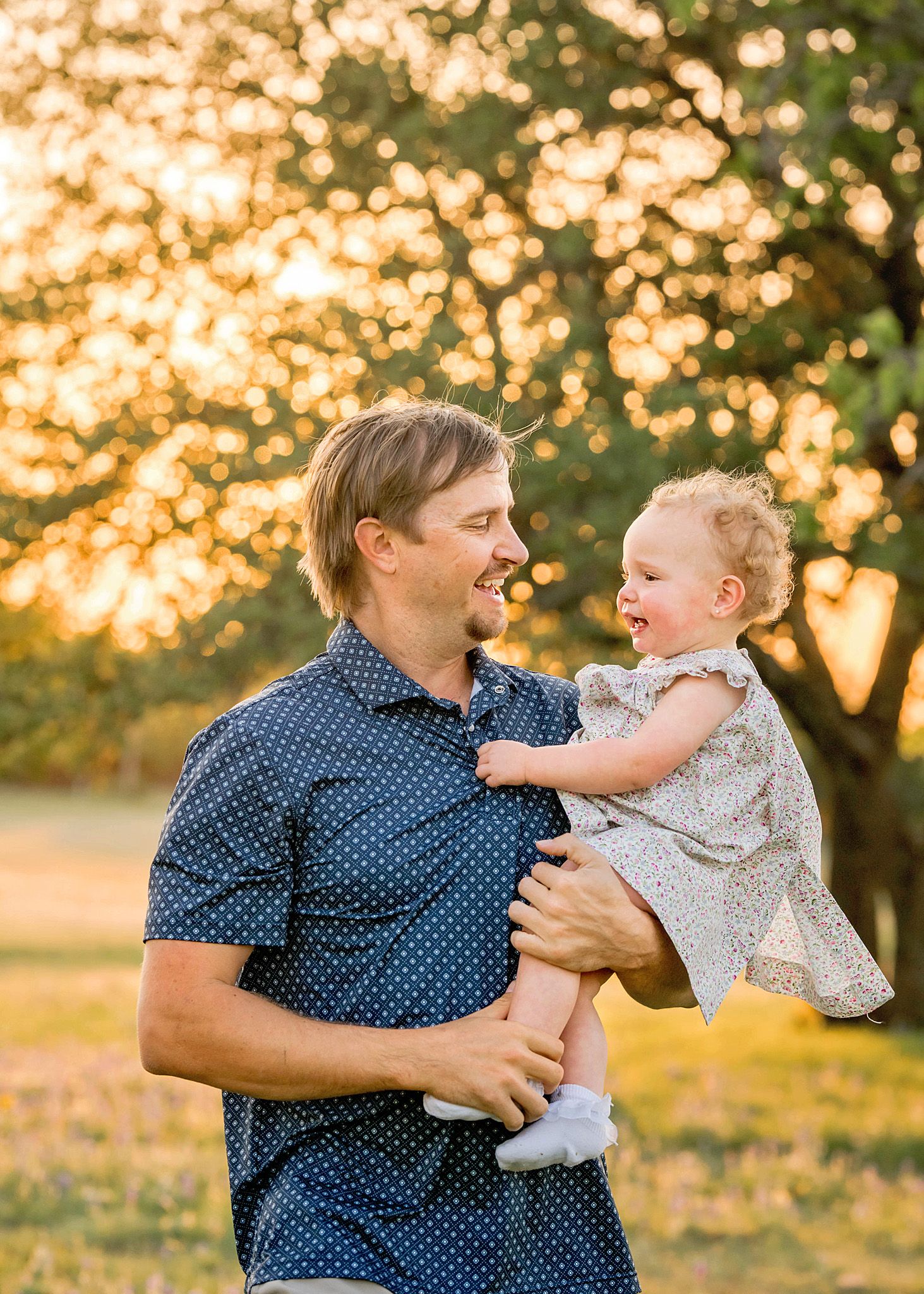 A smiling father holds his laughing toddler daughter outdoors during golden hour sunset light near a large tree.