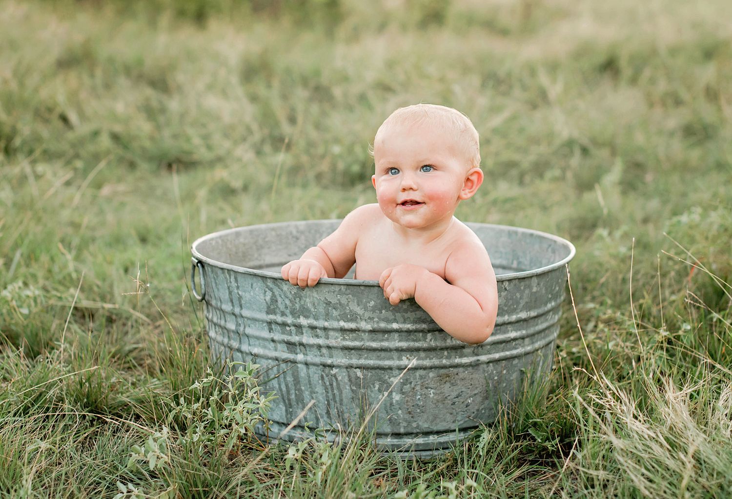 A happy baby sits in a rustic galvanized metal tub outdoors on grassy ground during a photo session.