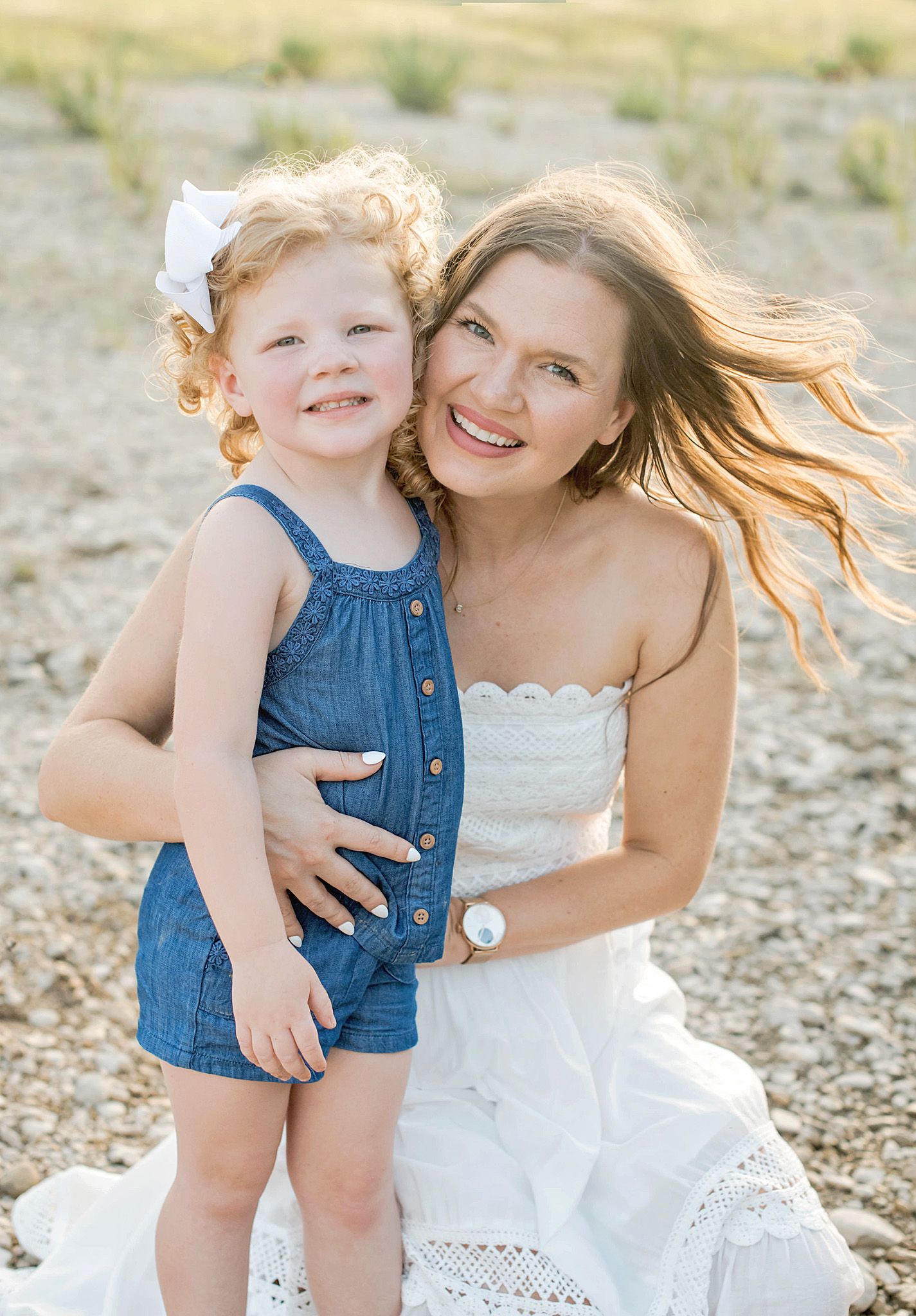 A smiling woman in a white dress hugs a curly-haired toddler girl in a denim romper outdoors.