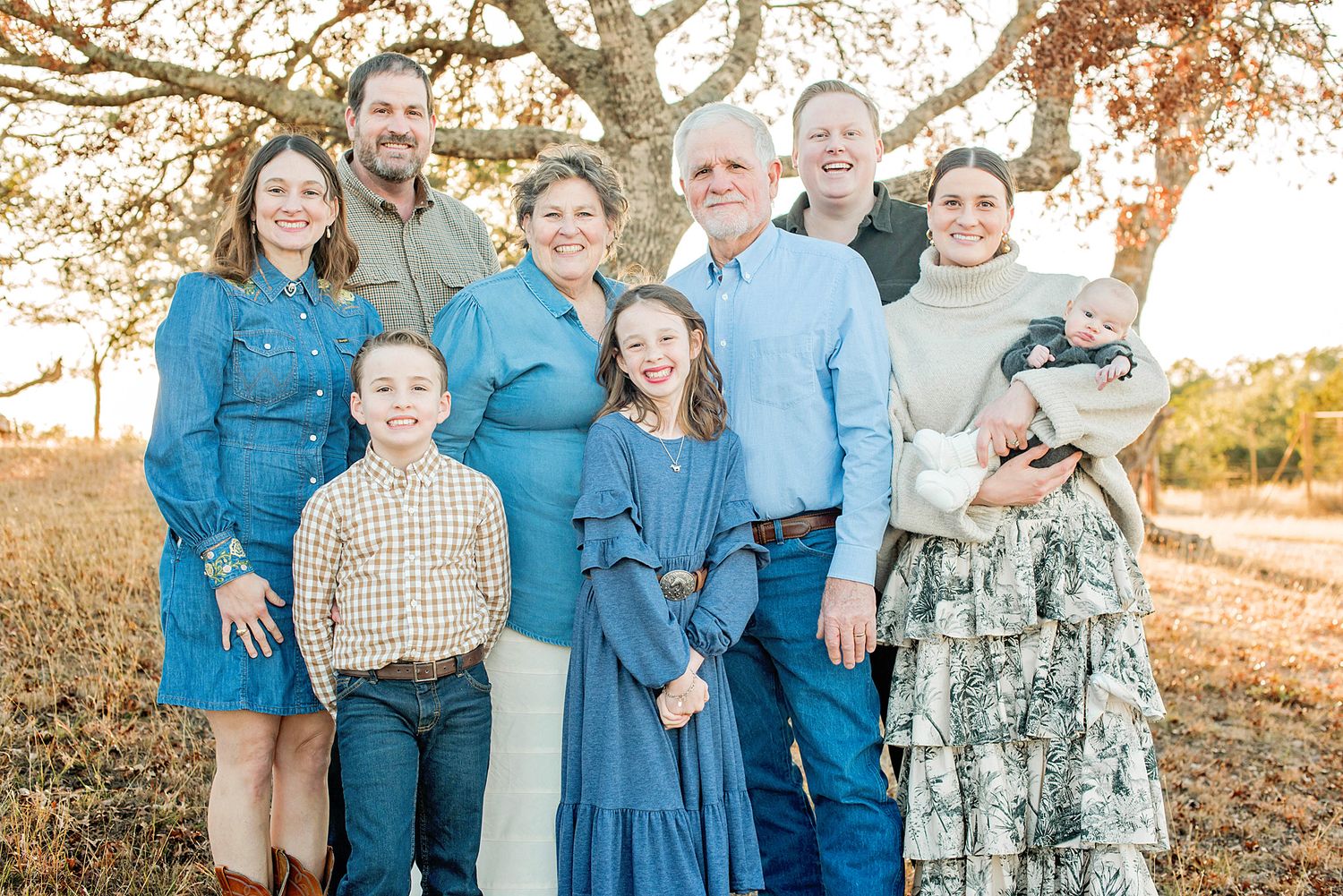 Multi-generational family portrait outdoors in autumn, smiling group of eight in coordinated blue outfits near a large tree.