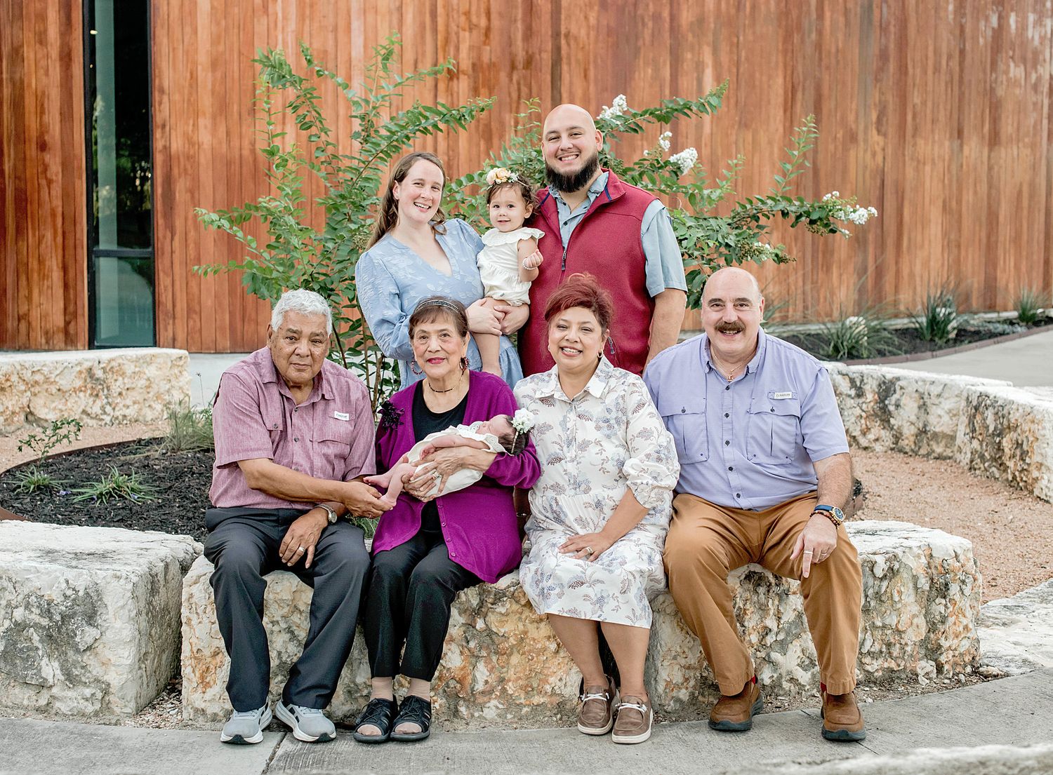 Multi-generational family portrait with seven members posing together outdoors near a stone wall and wooden building.