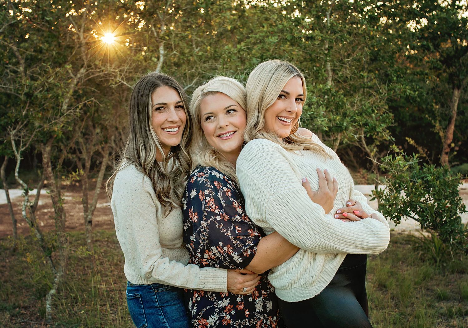 Three smiling women hugging outdoors at golden hour, surrounded by lush greenery in a warm, joyful portrait.