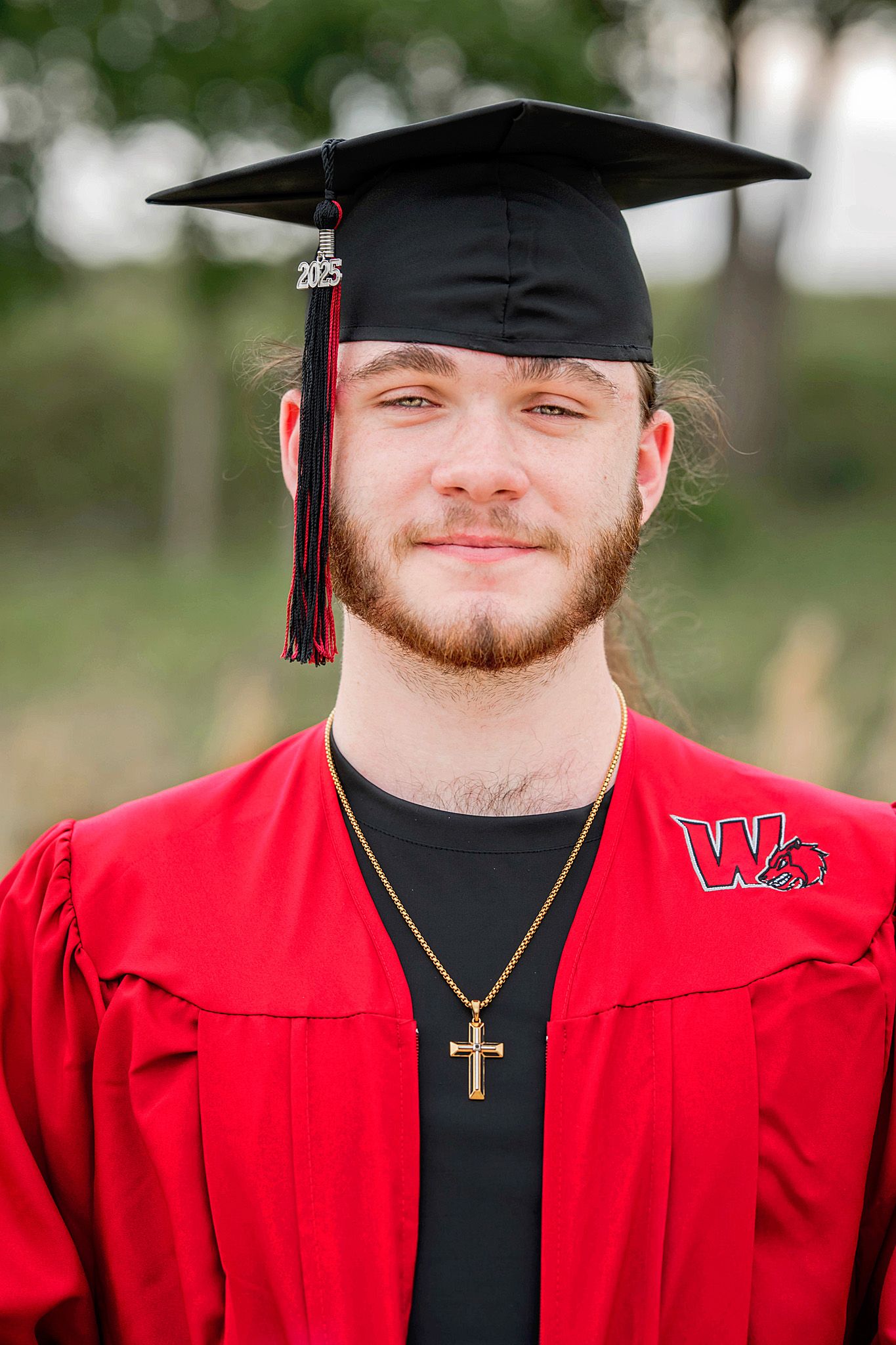 Young man in red graduation gown and 2025 cap wearing a cross necklace smiles outdoors at graduation.