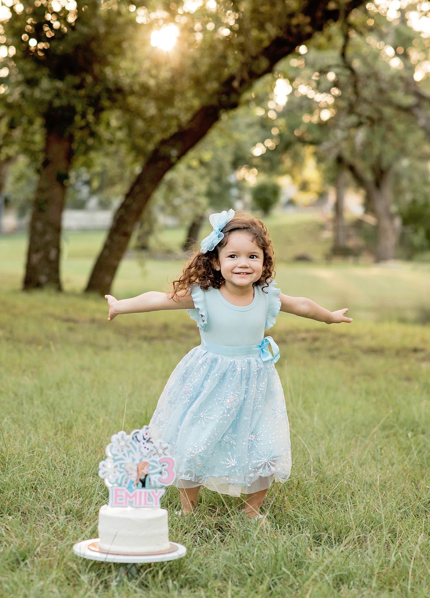A joyful toddler girl in a light blue lace dress spreads her arms wide near a birthday cake outdoors.