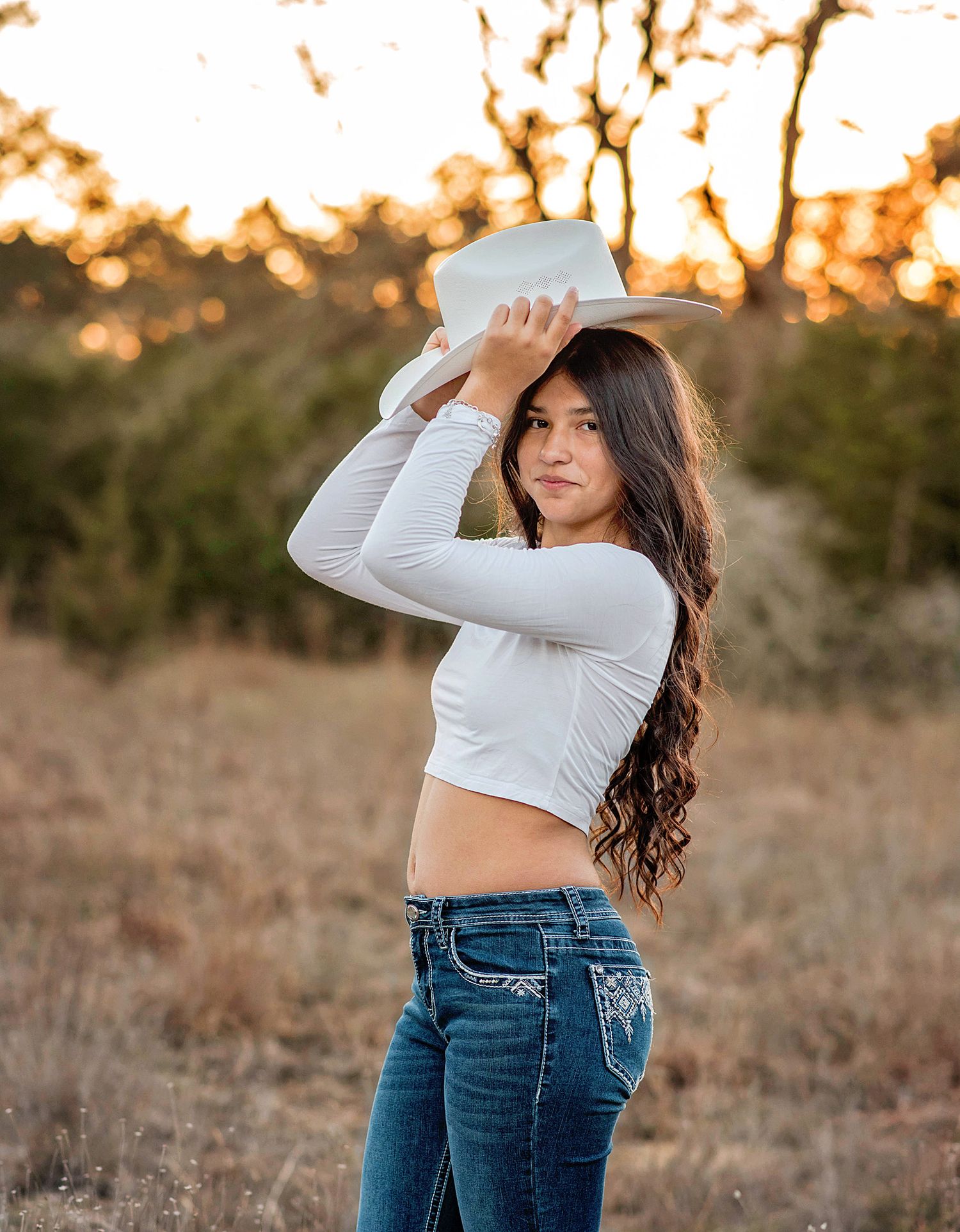 Young woman in white crop top and jeans holding a cowboy hat outdoors at golden hour.
