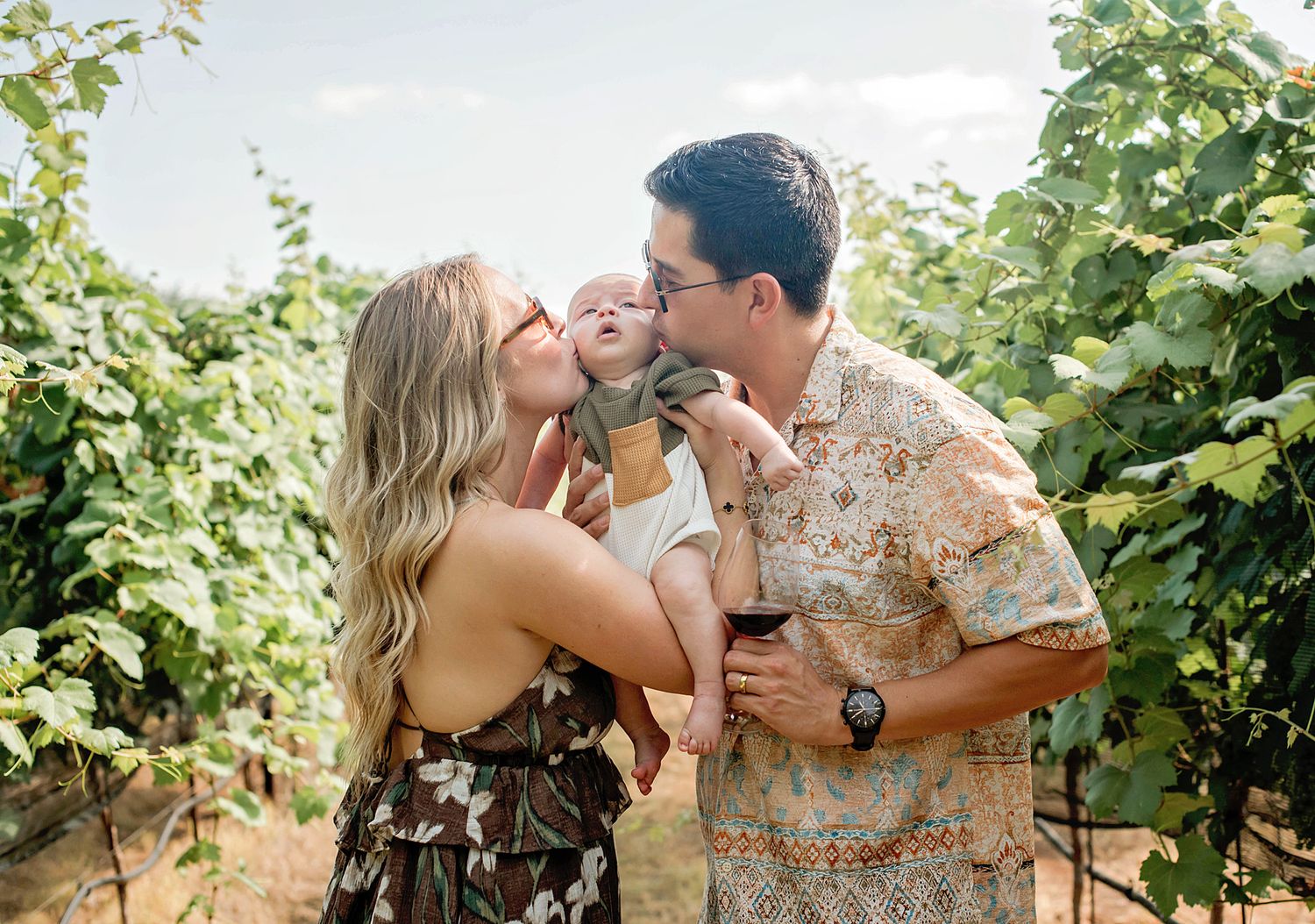 A couple kisses their baby at a sunny vineyard, with lush green grapevines in the background.
