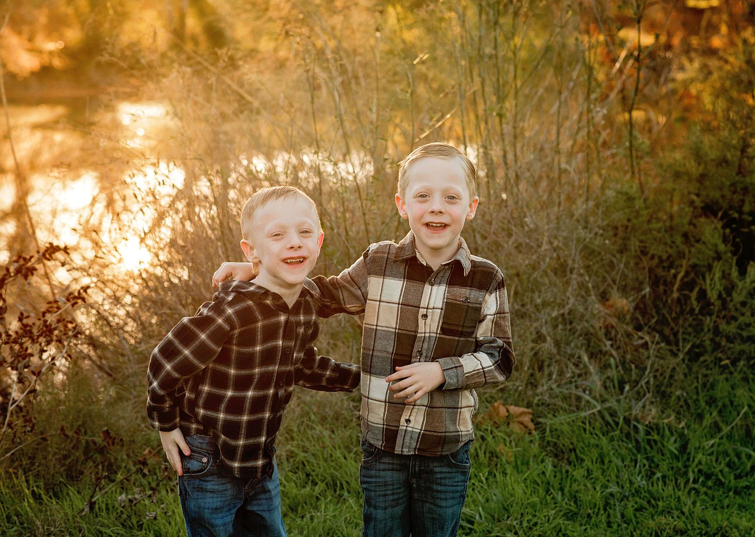 Two children in plaid shirts laugh and play together in a golden sunlit field at sunset.