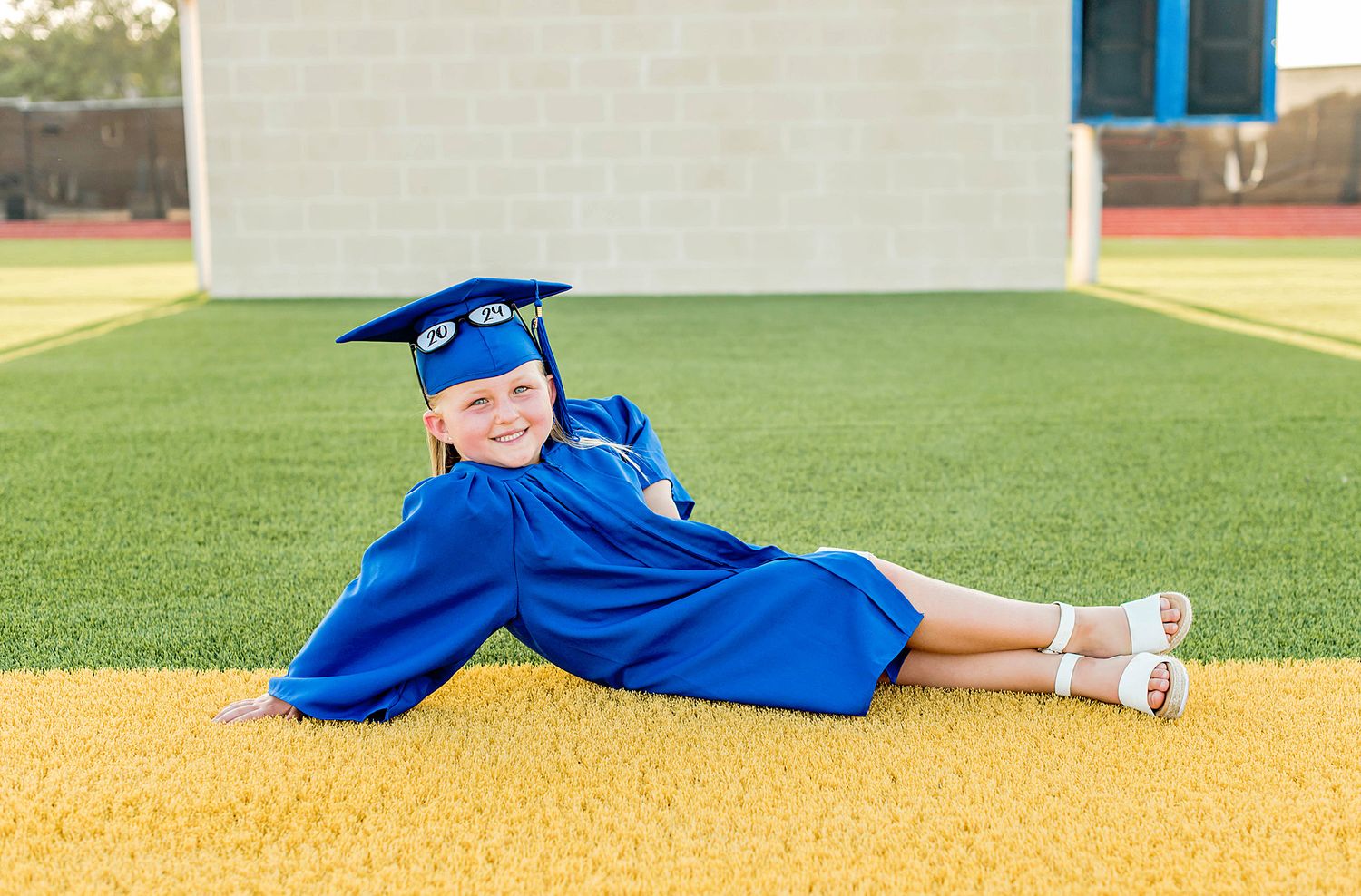 Graduate in blue cap and gown poses laying down on baseball field during graduation photo session.