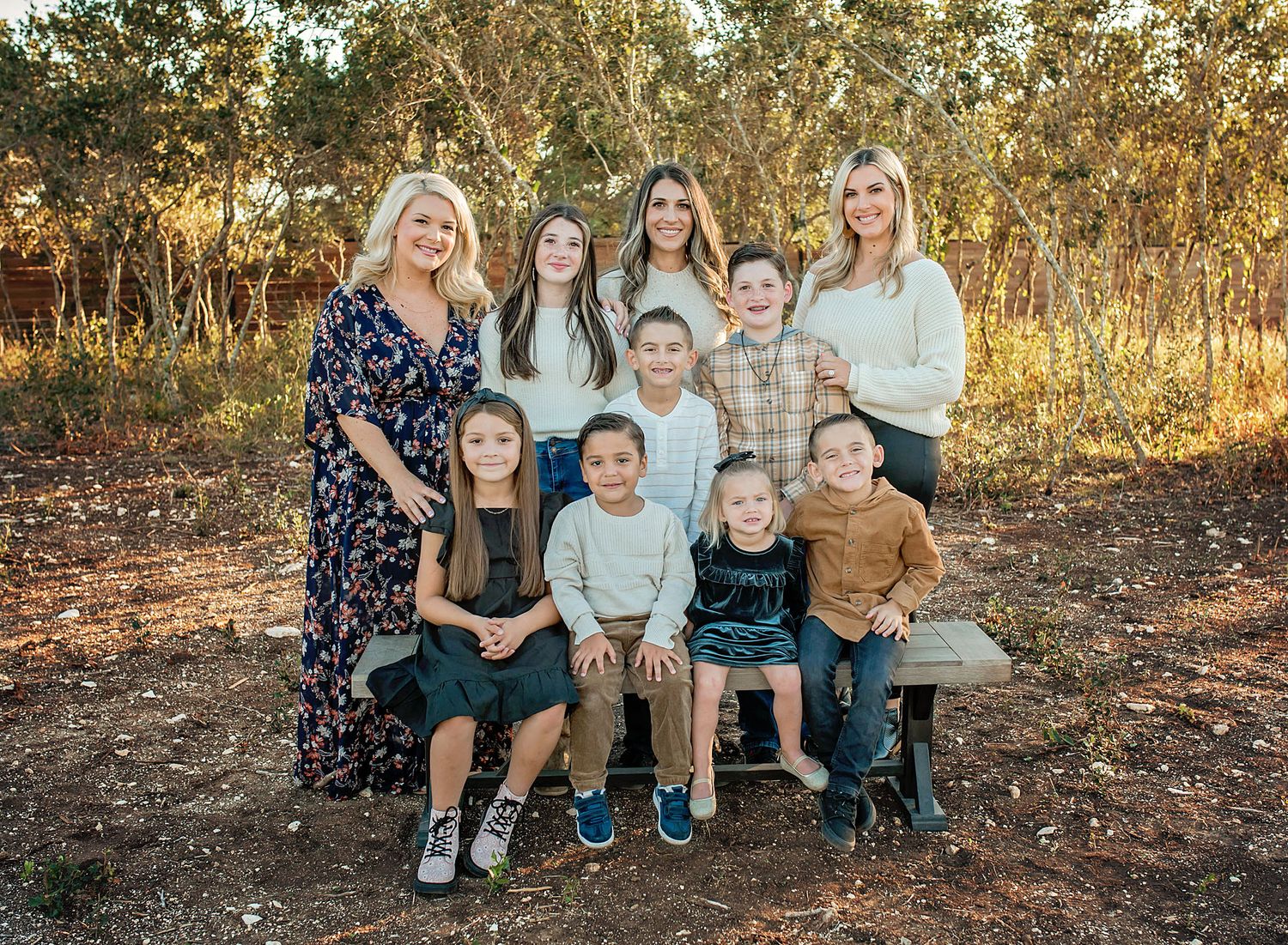 Family portrait of two women with seven children smiling outdoors on a bench surrounded by trees.