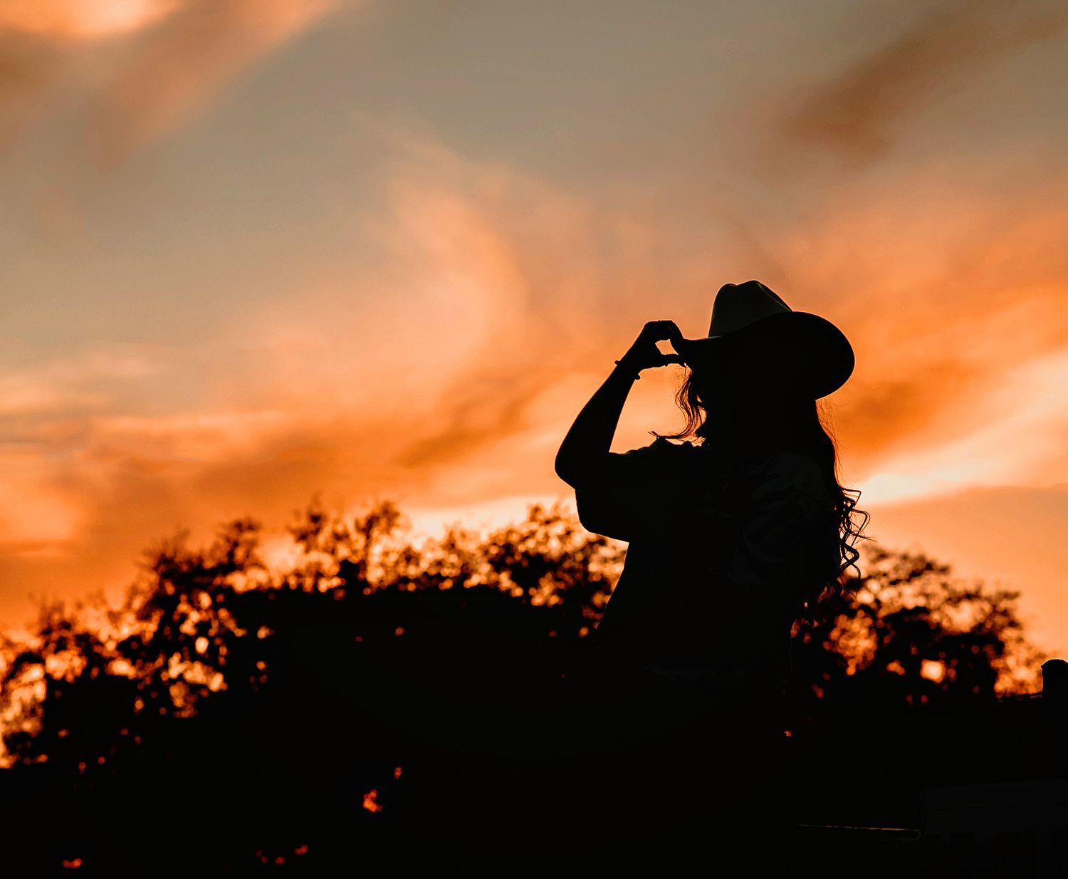 Silhouette of a woman in a cowboy hat posed against a dramatic orange sunset sky with dark trees.