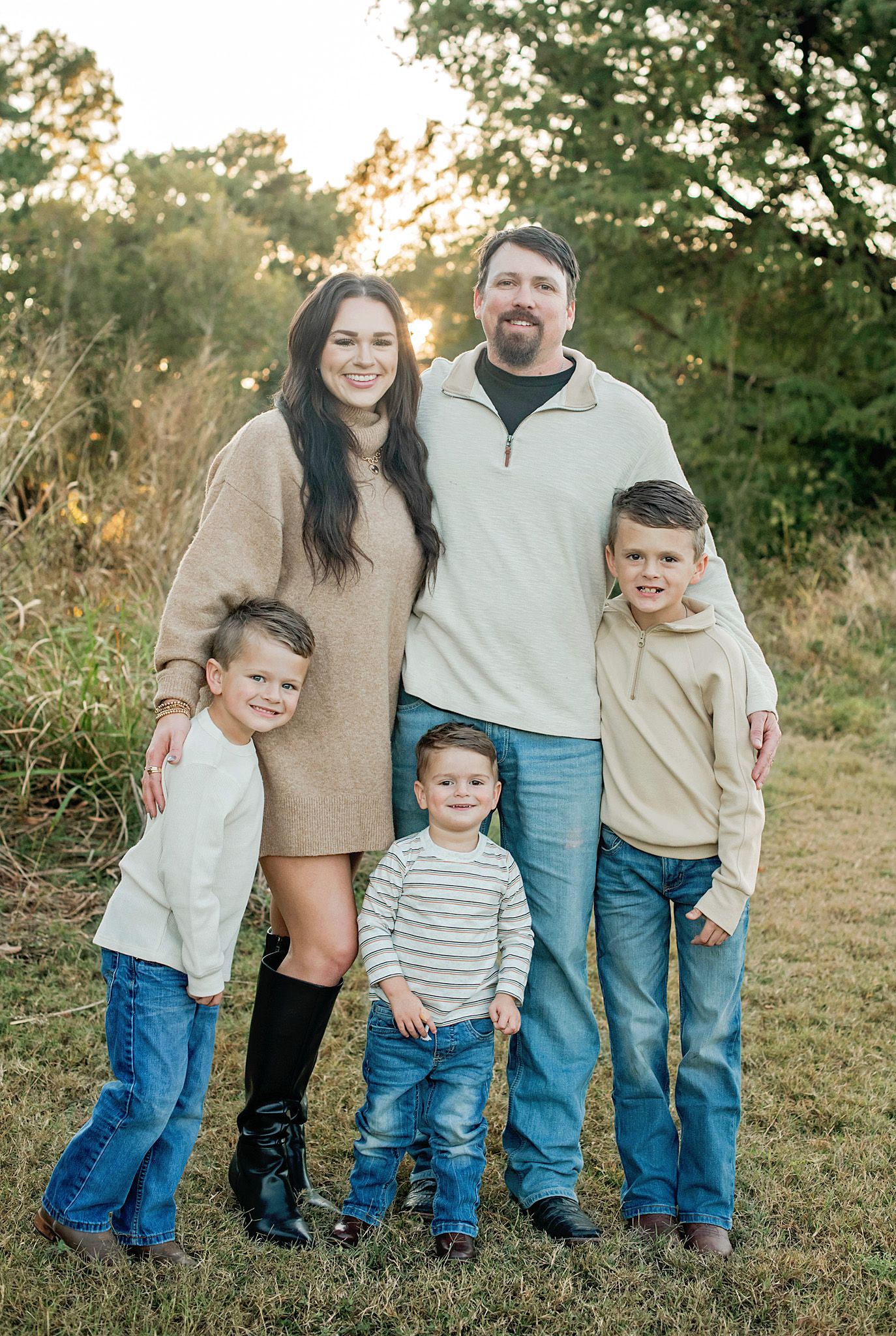 Family of five posing outdoors at golden hour, parents with three young boys in coordinating neutral outfits in a grassy field.
