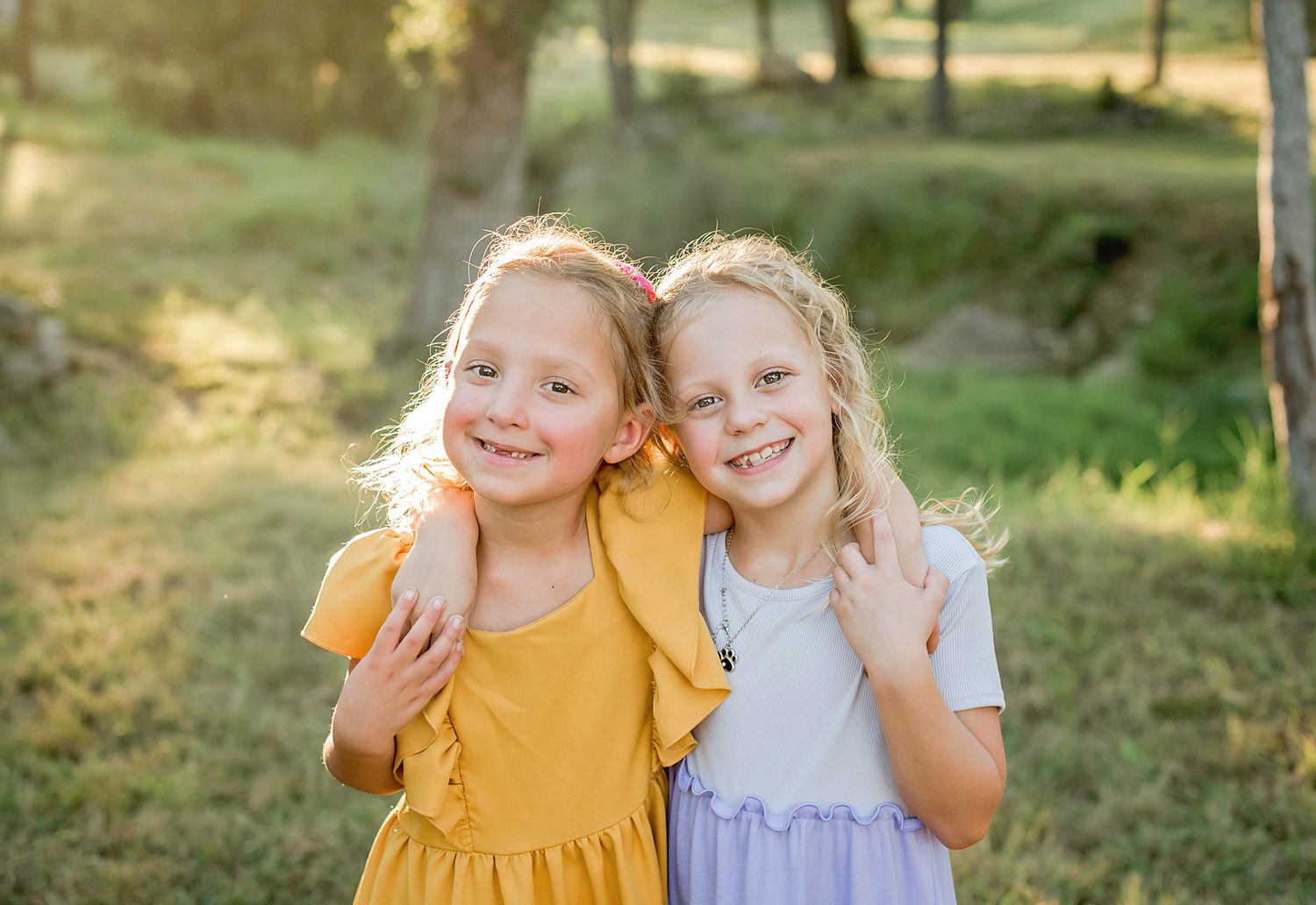 Two children in yellow and white dresses laugh and play together in a sunlit outdoor setting in Blanco, TX.
