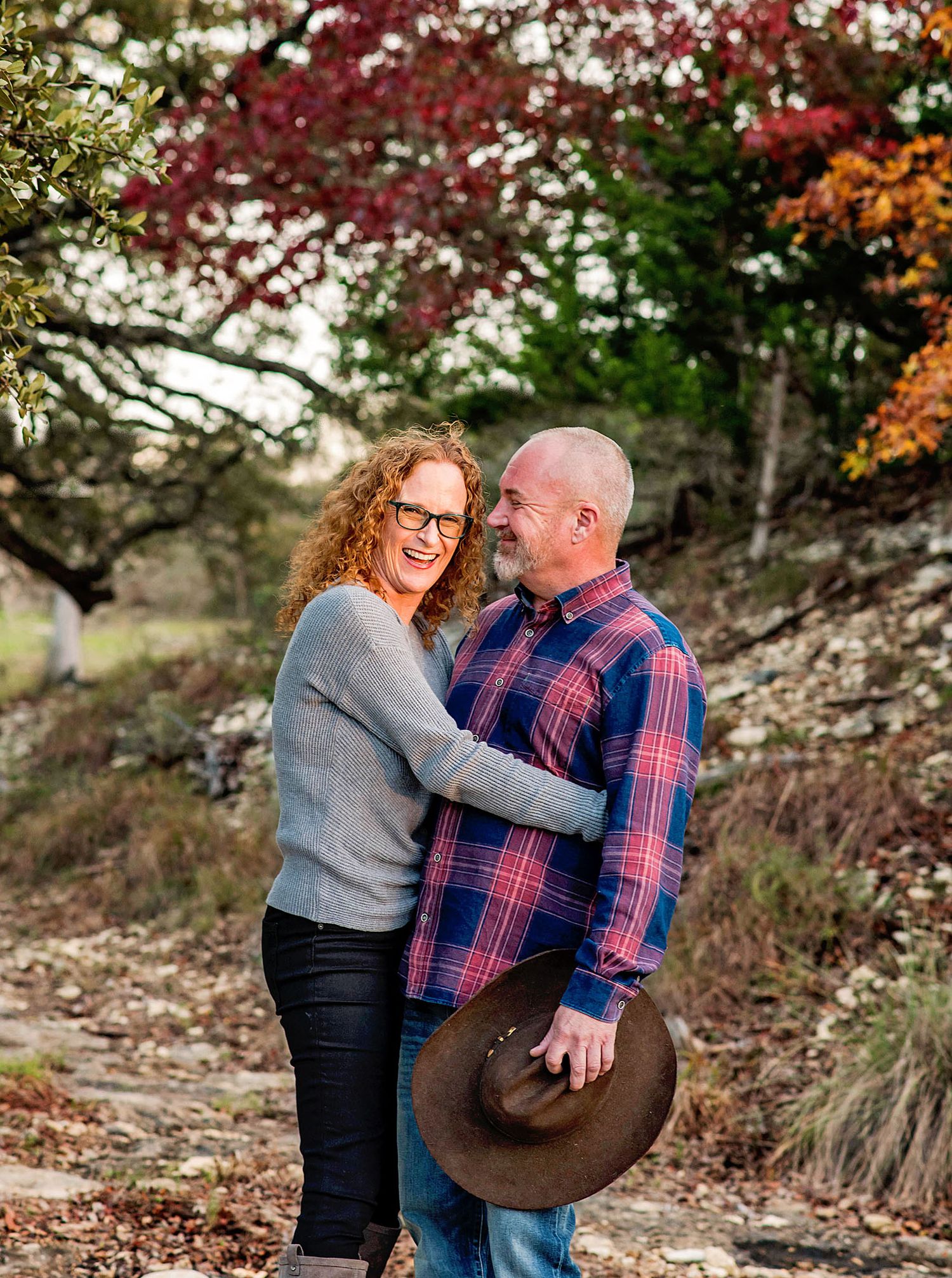 A couple shares a joyful moment together outdoors during an autumn photoshoot with colorful fall foliage in the background.