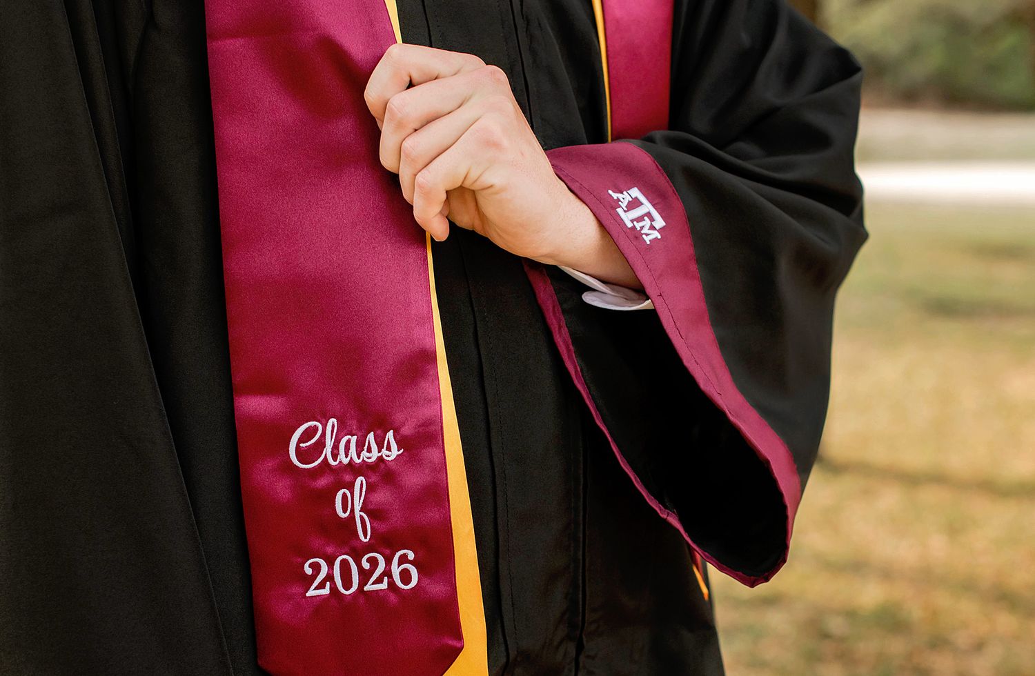 Graduate wearing black gown with maroon Class of 2026 stole, hands clasped during graduation ceremony.