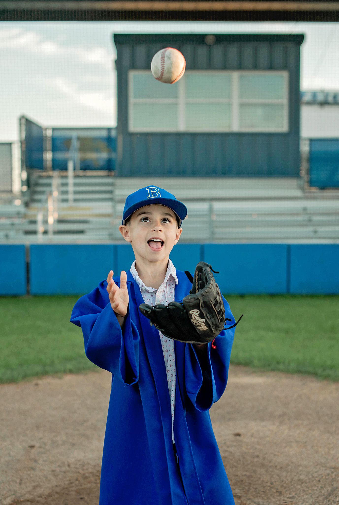 Young boy in blue graduation gown and cap catching a baseball with a mitt on a baseball field.
