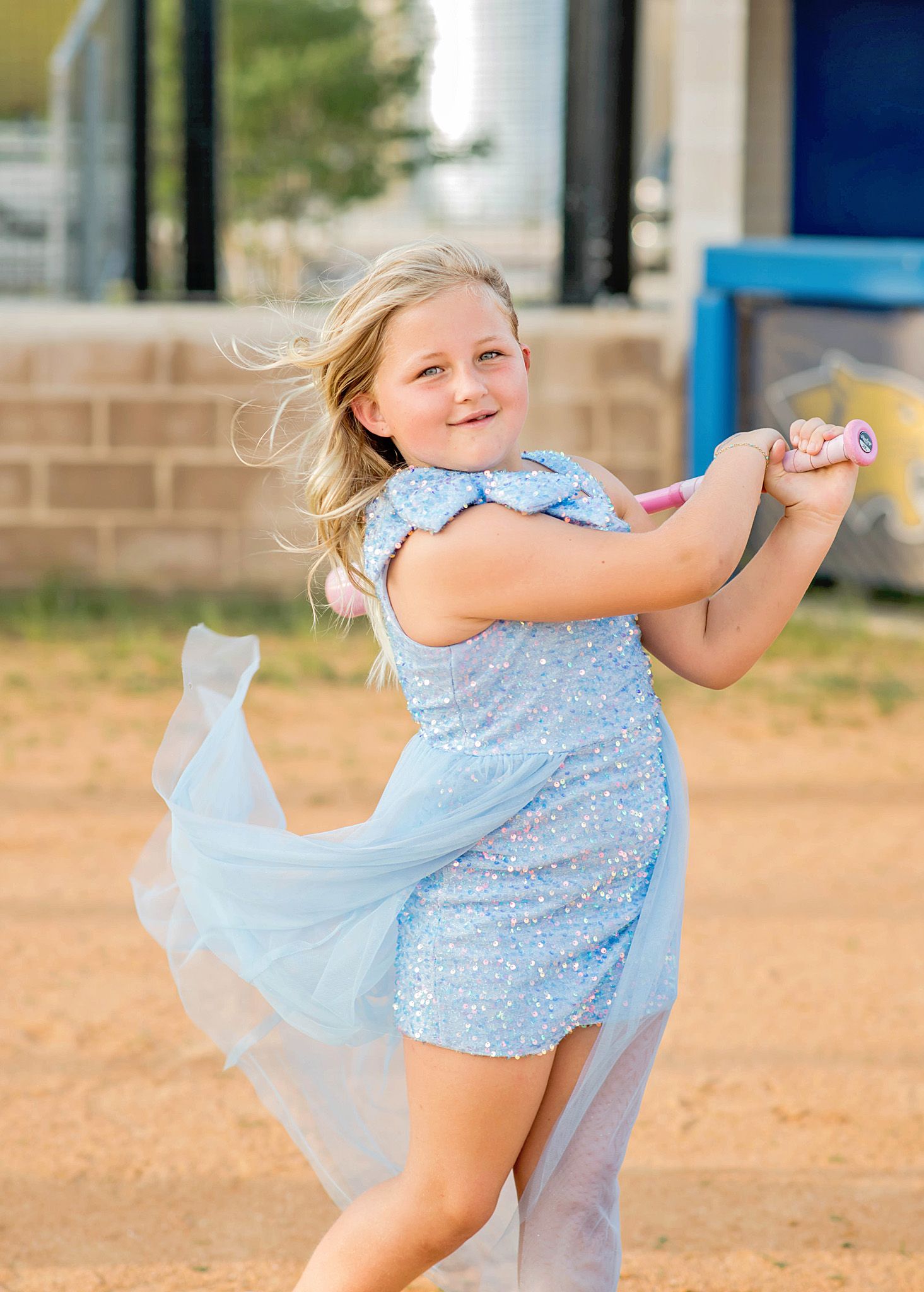 Young girl in a sparkling blue dress swings a pink bat on a dirt field, her hair and skirt flowing in the wind.