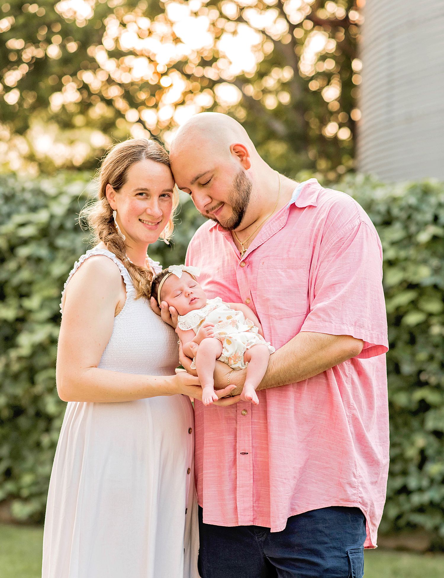 A couple smiles while cradling their newborn baby outdoors in golden hour light, the woman in white and man in pink.