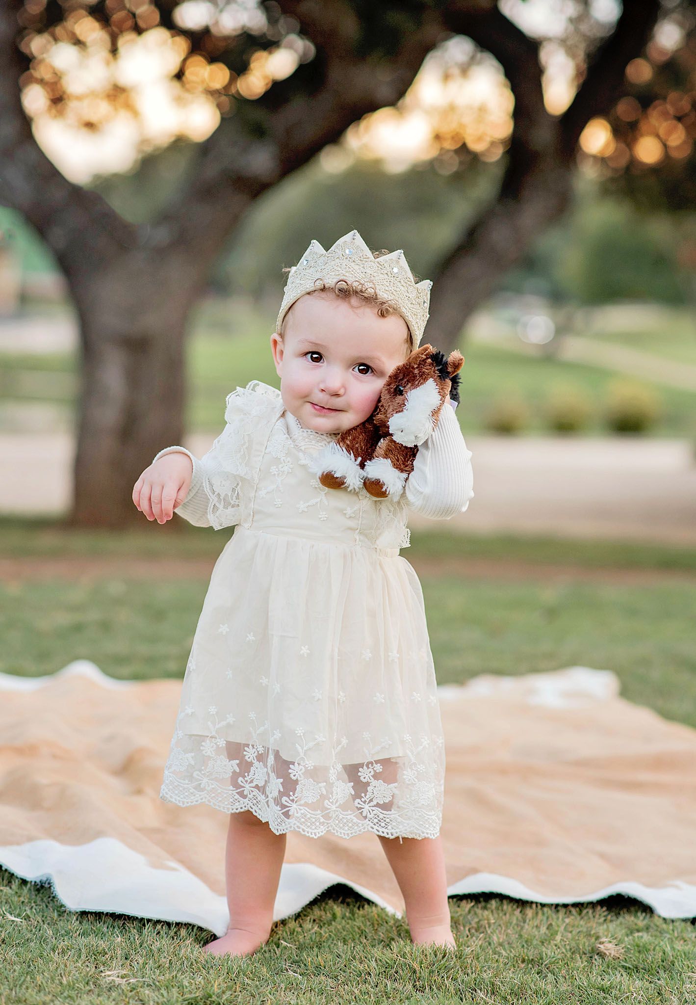 Toddler girl in ivory lace dress and crown holds stuffed animal outdoors on blanket with trees in background.