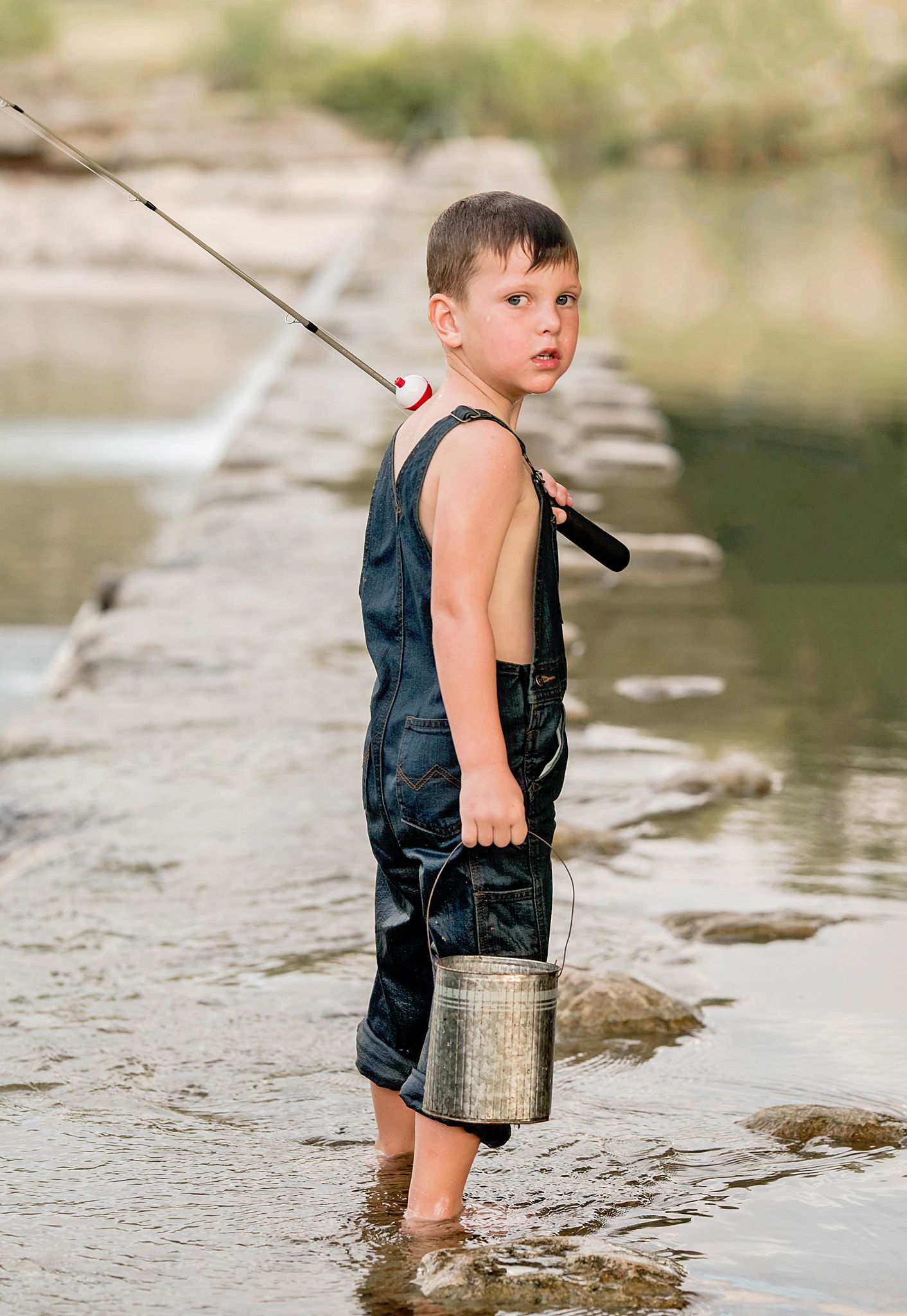 A young child wearing a navy tank top stands in shallow water while holding a fishing pole and metal bucket.