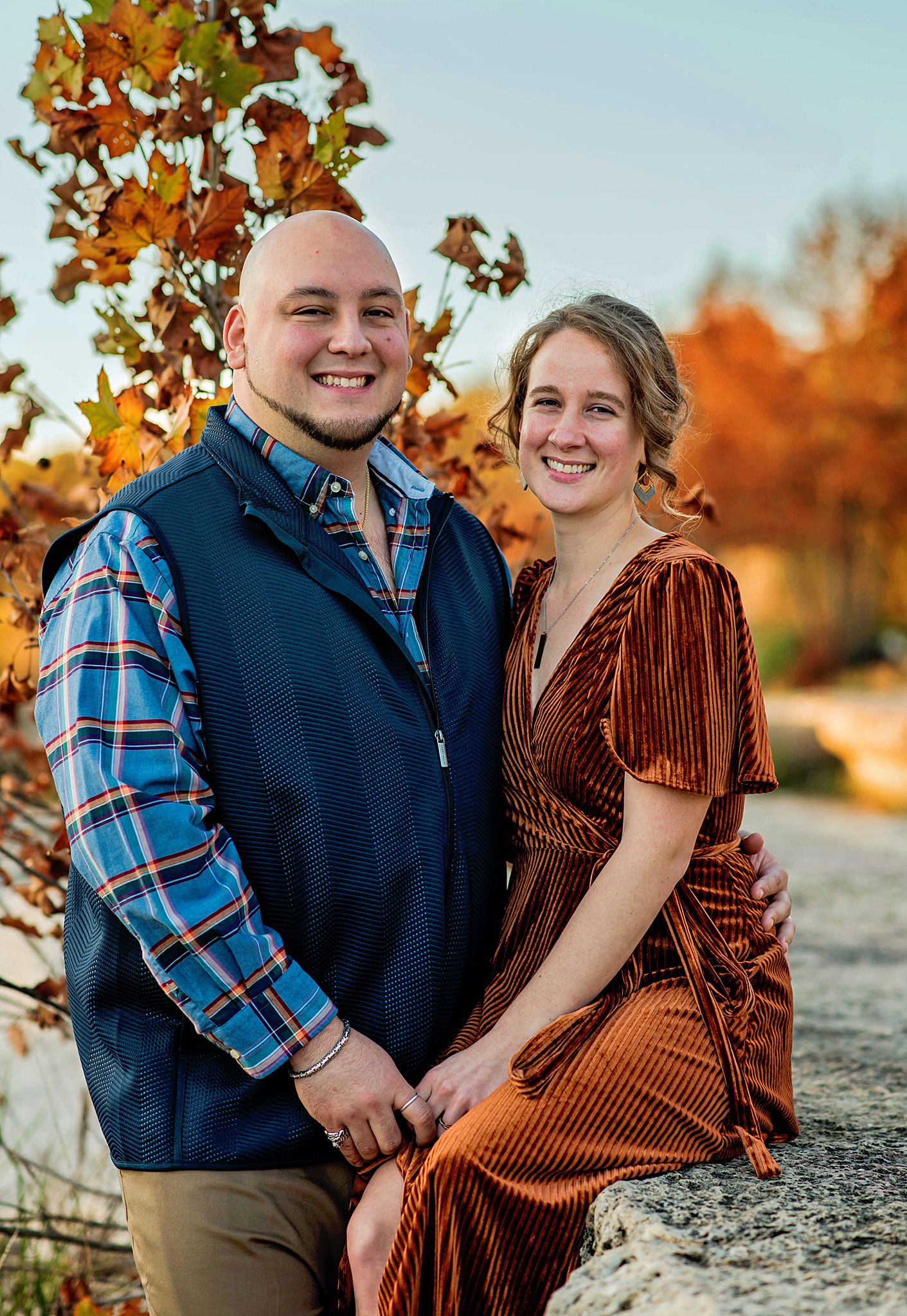 A couple embraces in front of autumn foliage wearing coordinated blue and rust colored outfits during a fall photoshoot.
