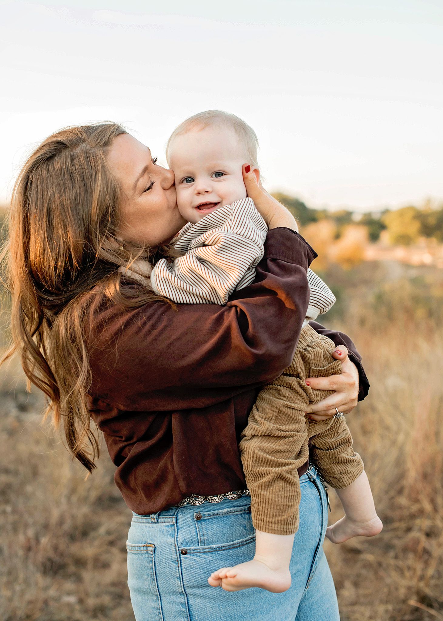 A parent in blue jeans and brown sweater holds and kisses a baby wearing beige knit clothing in a golden autumn field.
