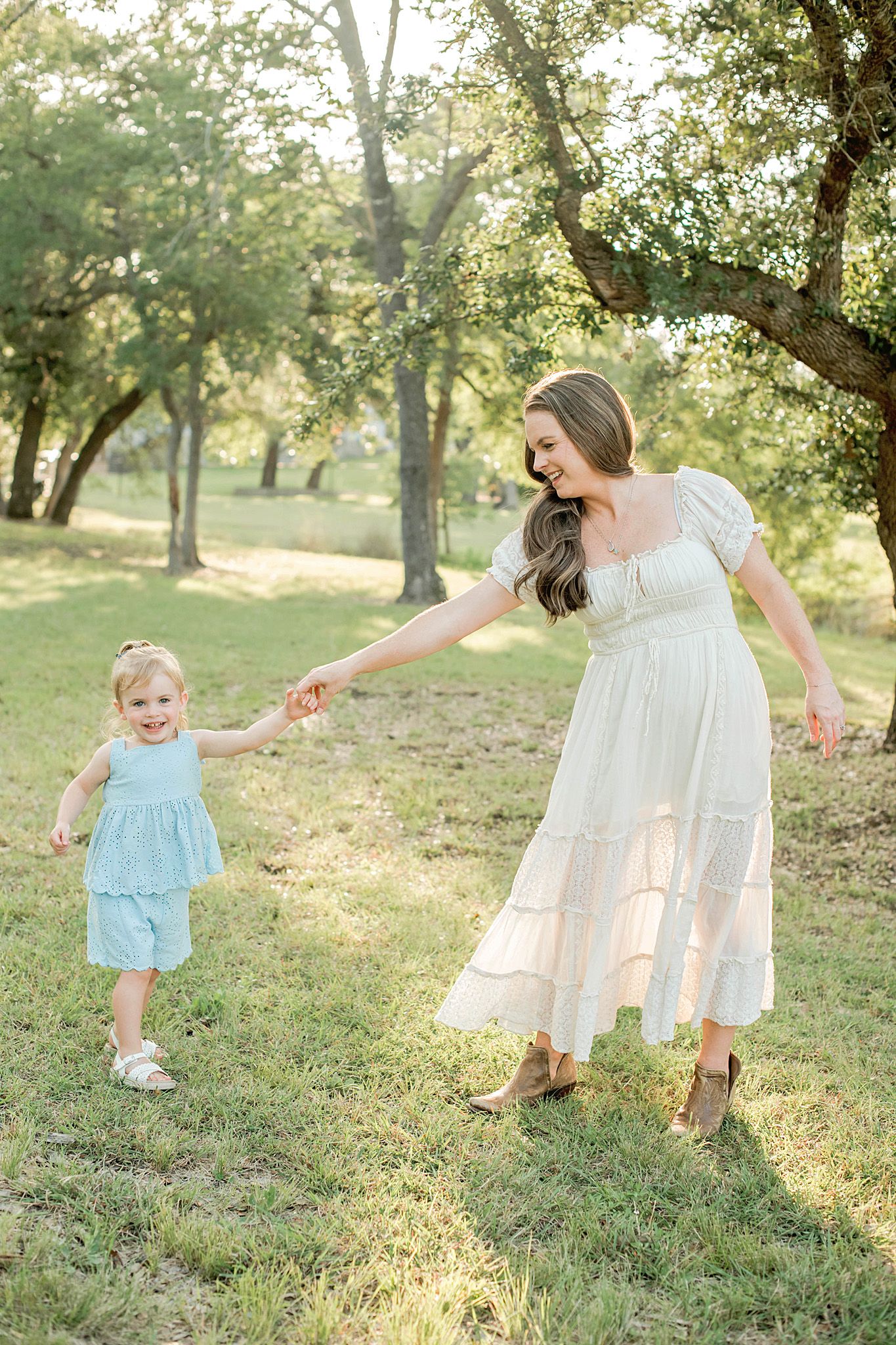 A mother in a white dress holds hands with her toddler in a light blue outfit while walking in a sunny green park.