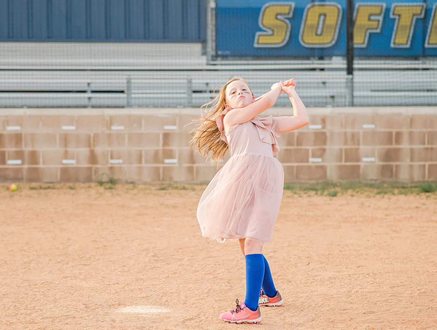 Young girl in pink dress and blue knee socks swinging a bat on a softball field.