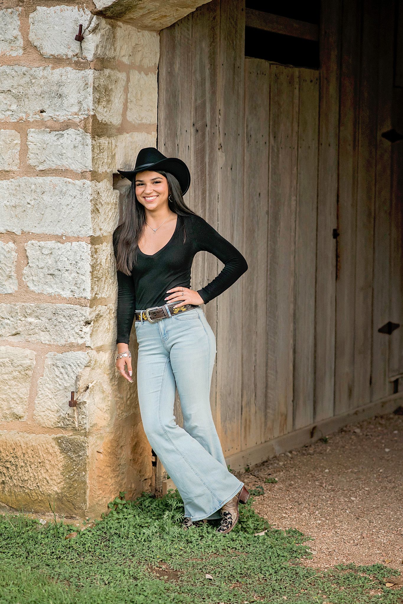 Woman in black cowboy hat, black top, and flare jeans leaning against a rustic stone barn wall.