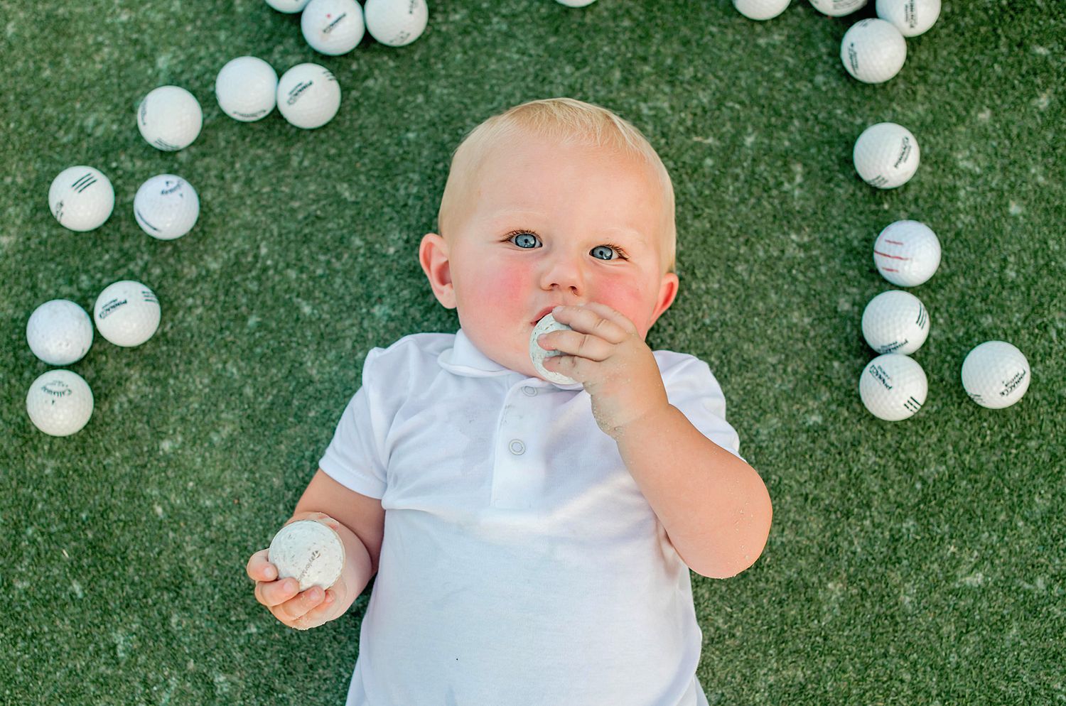 Baby lying on green grass surrounded by white golf balls looking up at the camera.