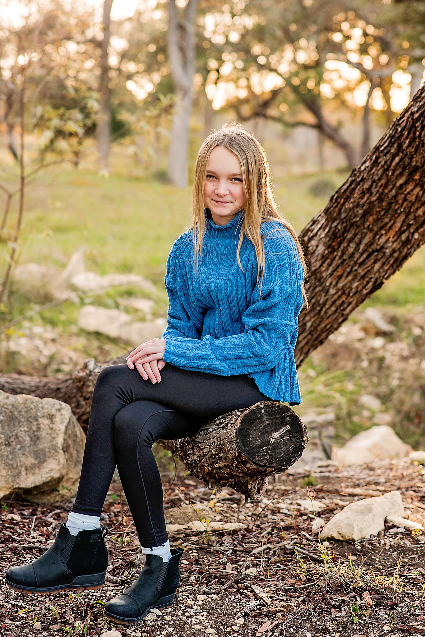 Young person in blue sweater sits on tree trunk in natural woodland setting.