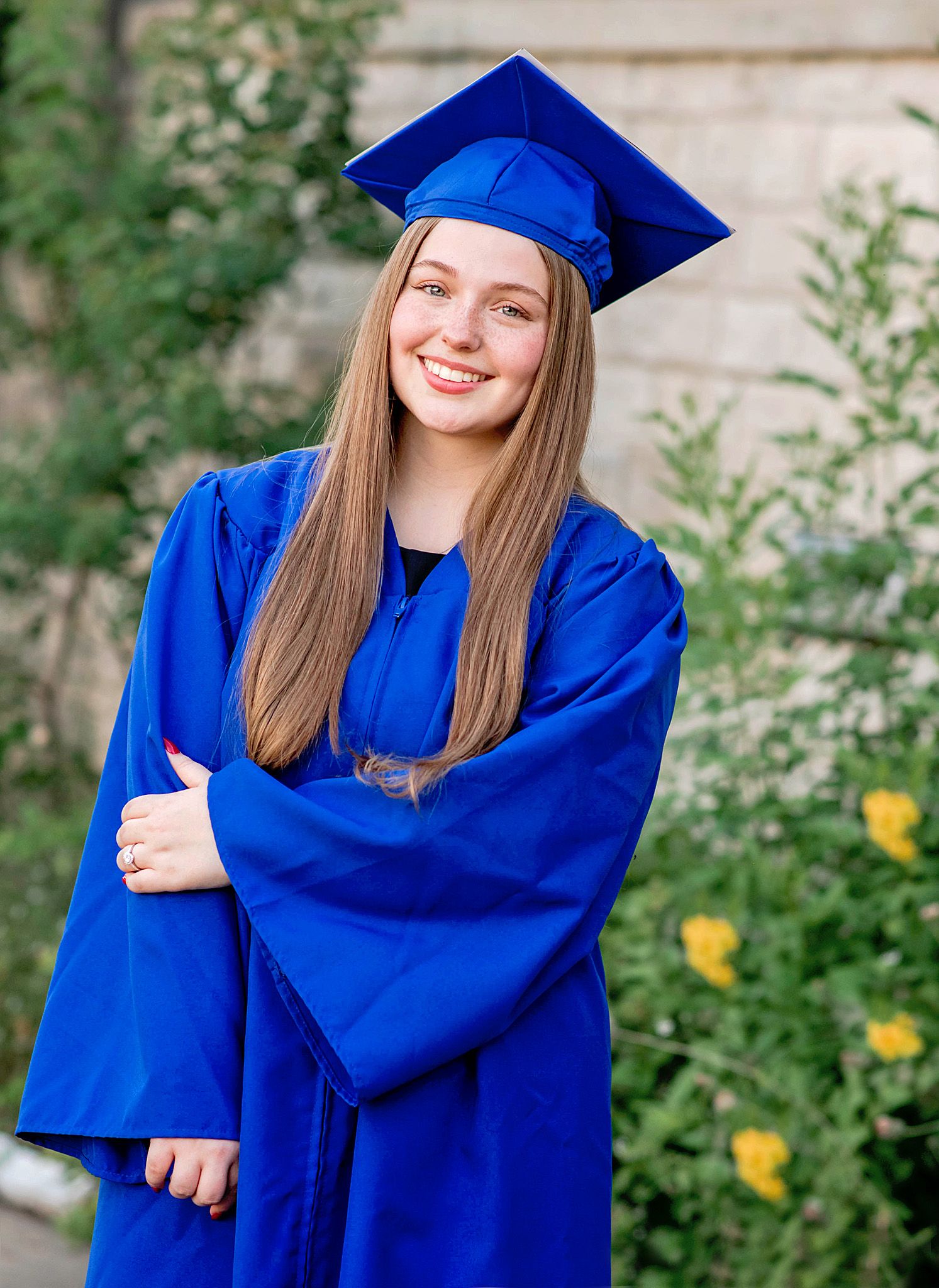 Smiling graduate in blue cap and gown posing outdoors with arms crossed in front of green foliage.