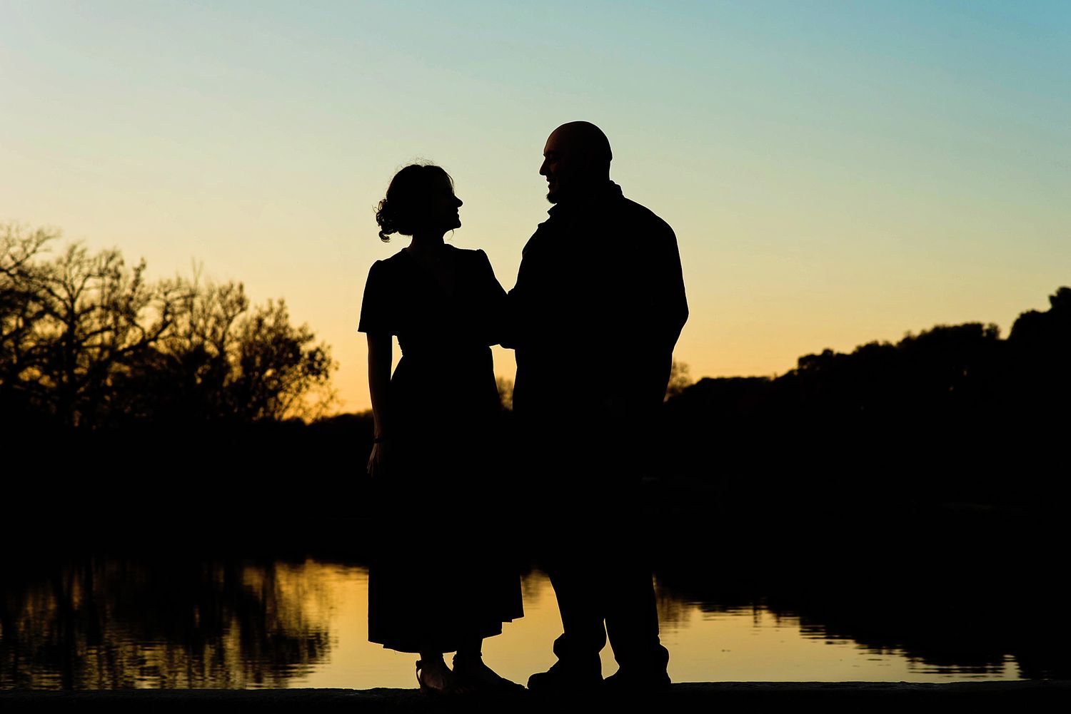 Silhouettes of people against a sunset sky reflected in calm water at dusk.
