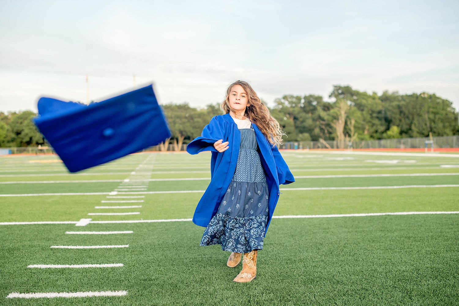 A joyful graduate tosses her blue cap on a green football field, wearing a blue gown and floral dress.