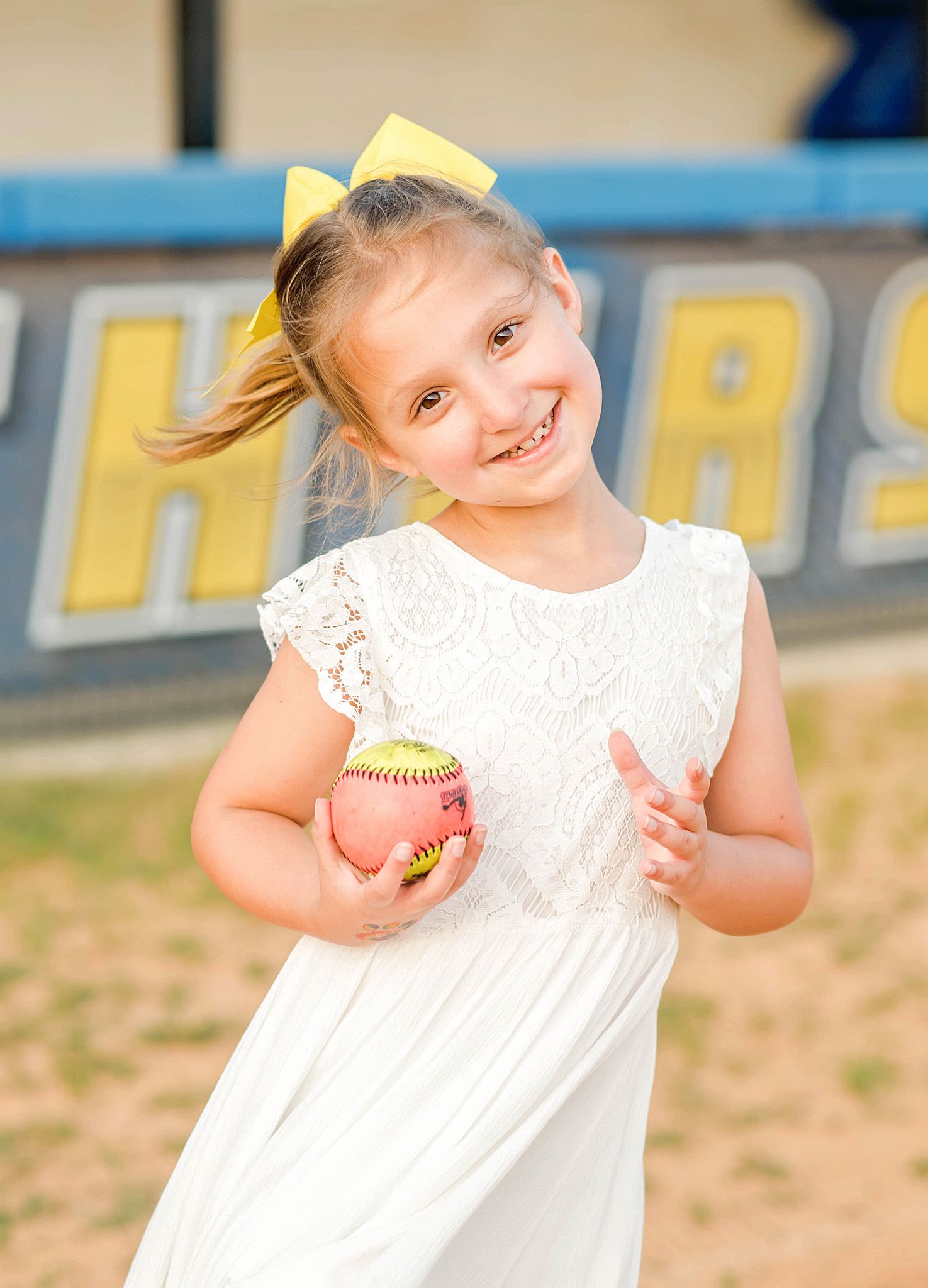 Young girl in white dress holding a softball, smiling on a baseball field with yellow bow in hair.