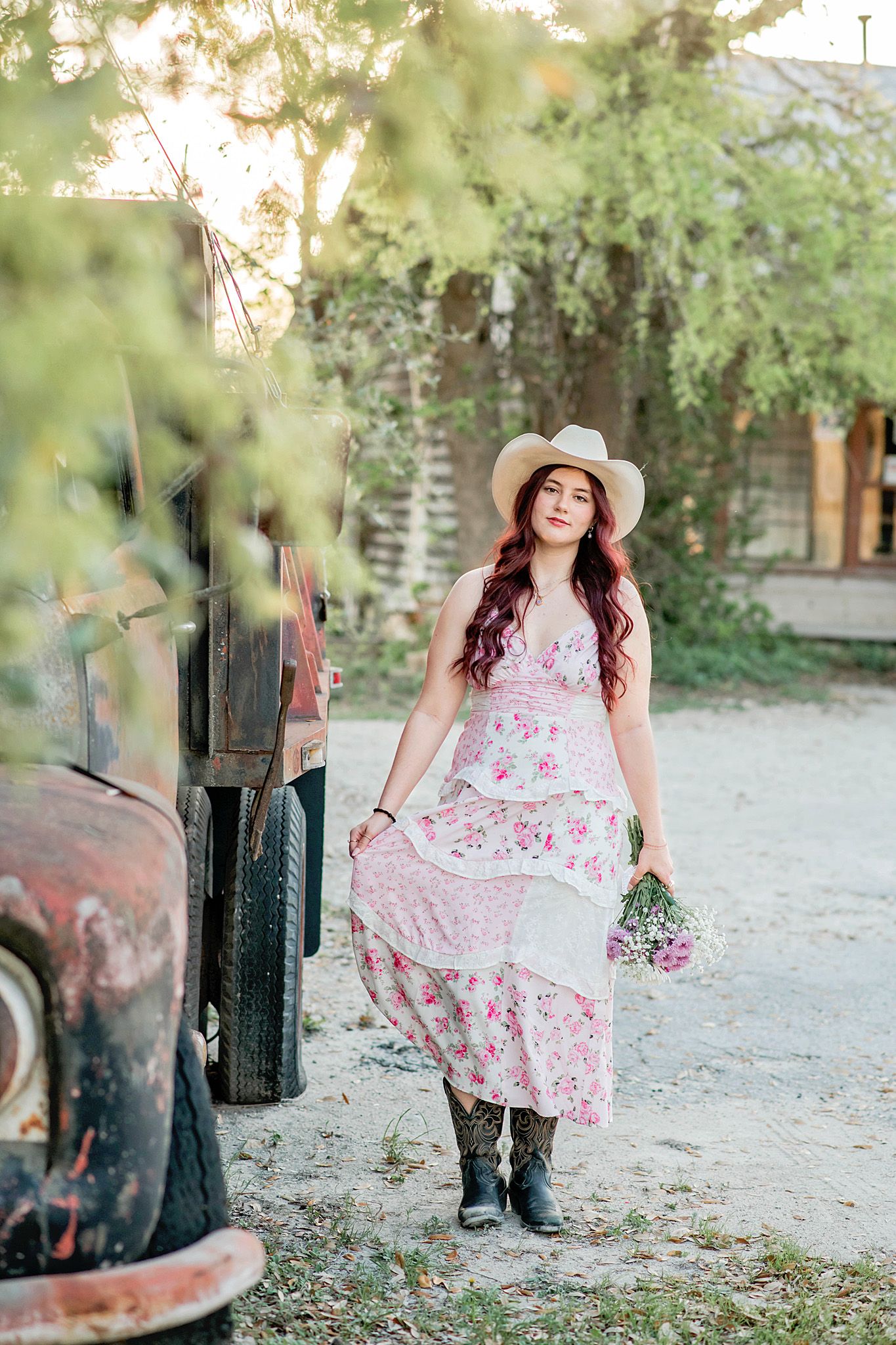 Young woman in floral dress, cowboy hat, and boots holding wildflowers beside a vintage rusty truck outdoors.