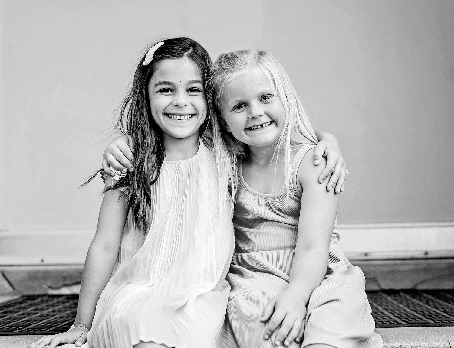 Two smiling young girls in dresses pose together in a black and white portrait photo.