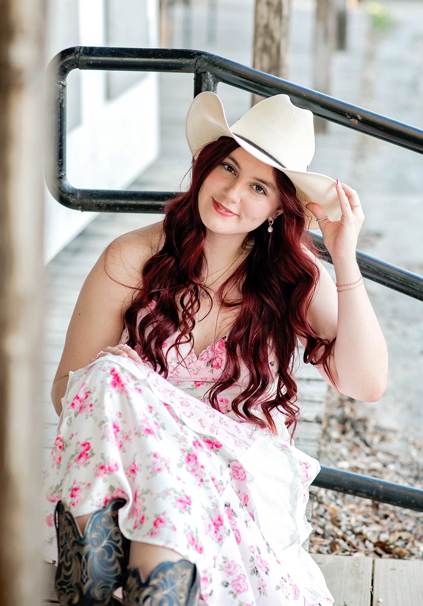 Young woman with red hair wearing a white cowboy hat, floral dress, and cowboy boots sitting on outdoor steps.