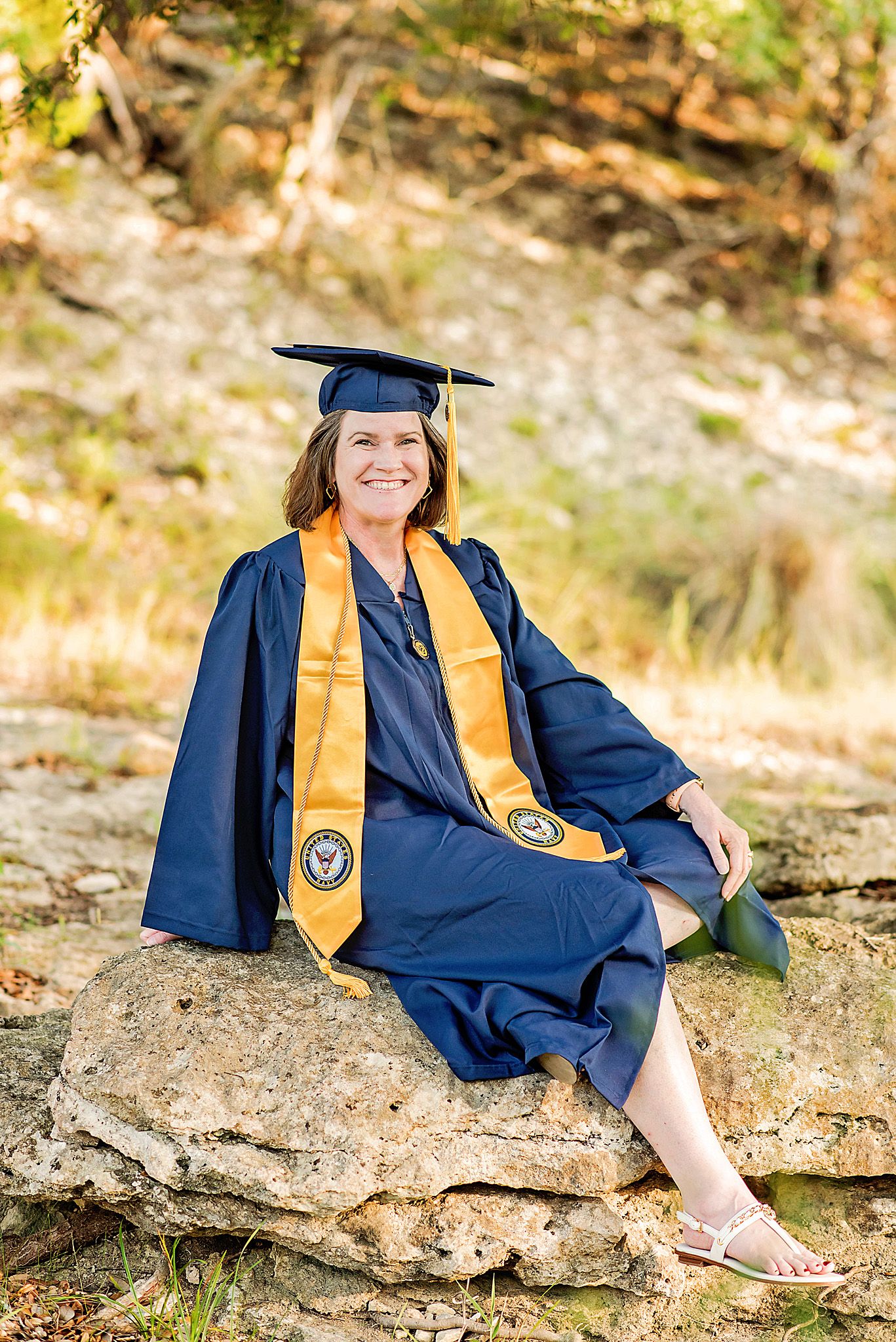 Woman in navy graduation gown and gold stole sits on rocks outdoors, smiling proudly in cap and gown.