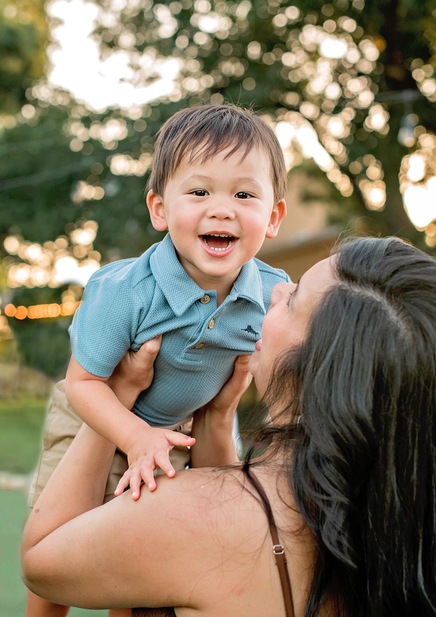 A joyful toddler in a blue polo shirt laughs while being lifted by a woman outdoors in warm golden light.