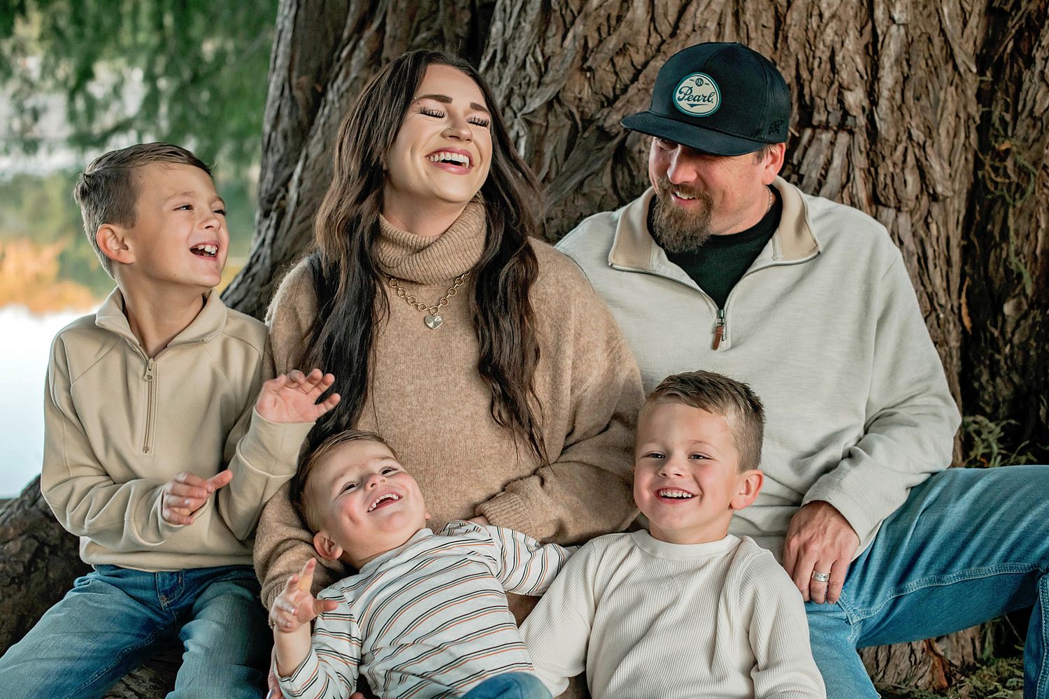 Happy family of five laughing together outdoors, sitting by a large tree in a cozy autumn setting.