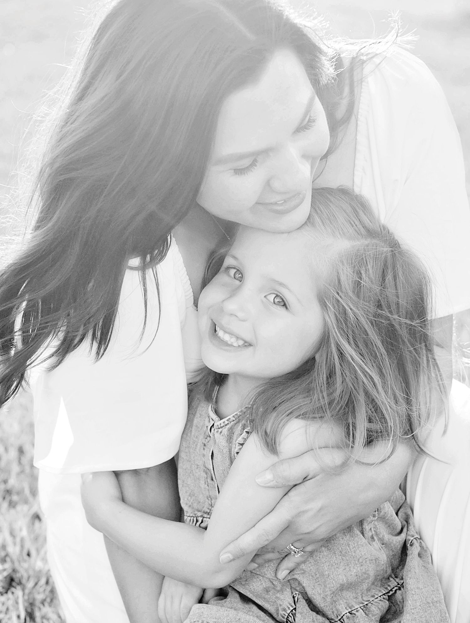 Black and white photo of a mother tenderly embracing her smiling young daughter outdoors.