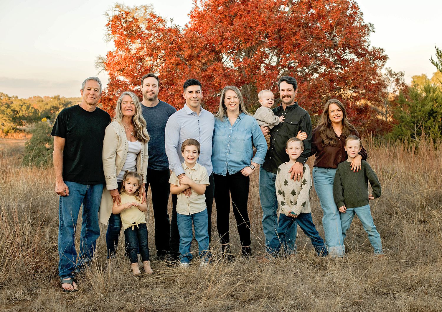 Large family group portrait outdoors in autumn field with red foliage tree backdrop during golden hour.