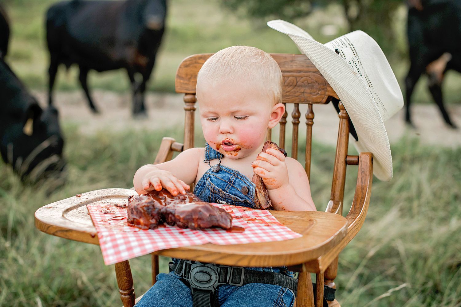 A small child in a wooden high chair enjoys bbq ribs on a red checkered tablecloth with a cowboy hat resting nearby.