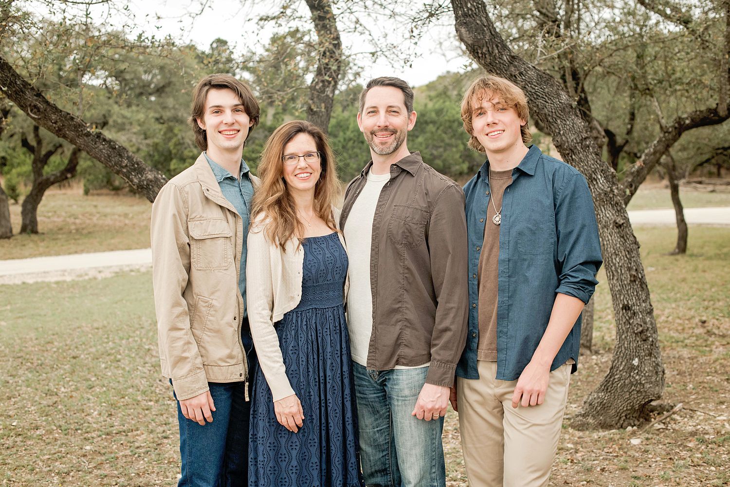 Family of four smiling outdoors among trees, featuring two teenage boys, a woman, and a man in casual attire.