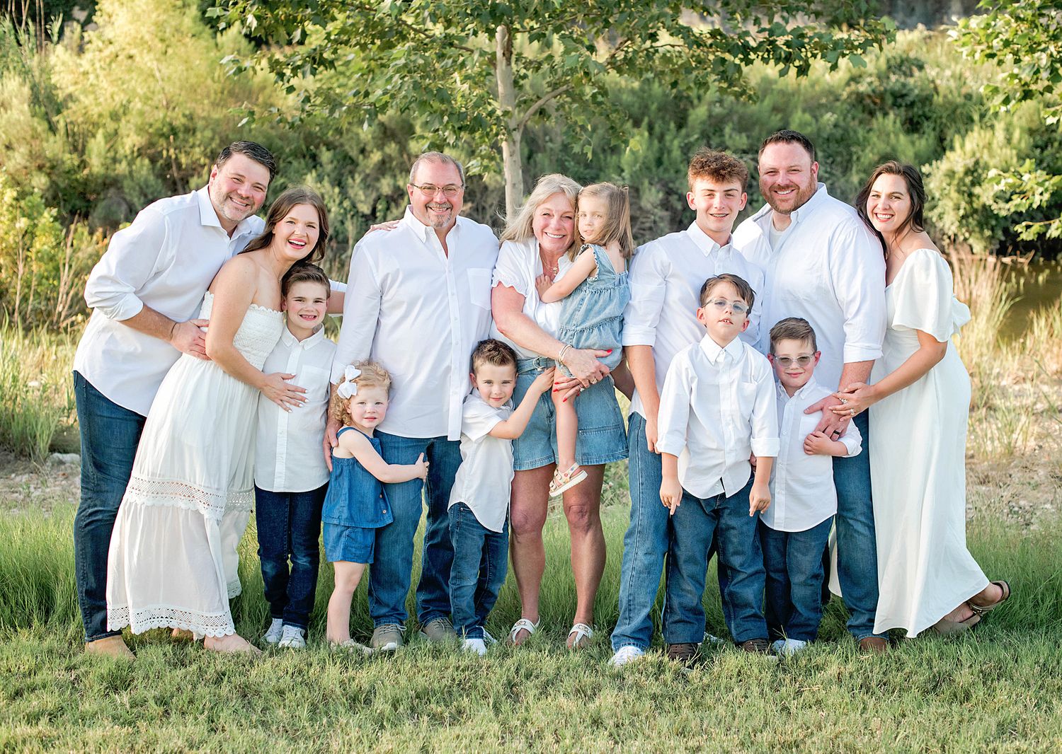 Large family portrait outdoors in white and blue outfits, smiling together on green grass with trees in background.