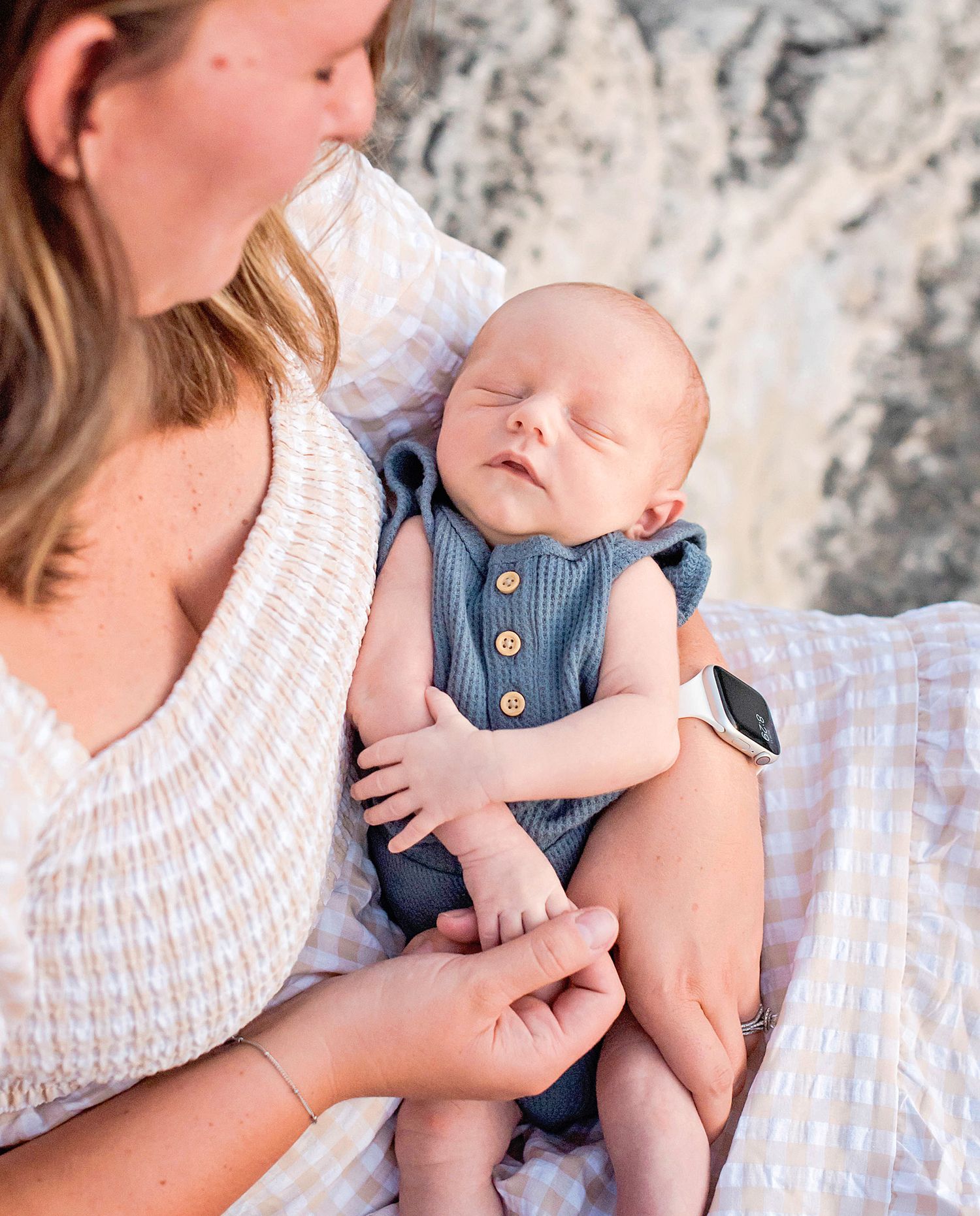 A sleeping newborn baby wearing a blue romper rests peacefully in a caregiver's arms against a textured gray background.